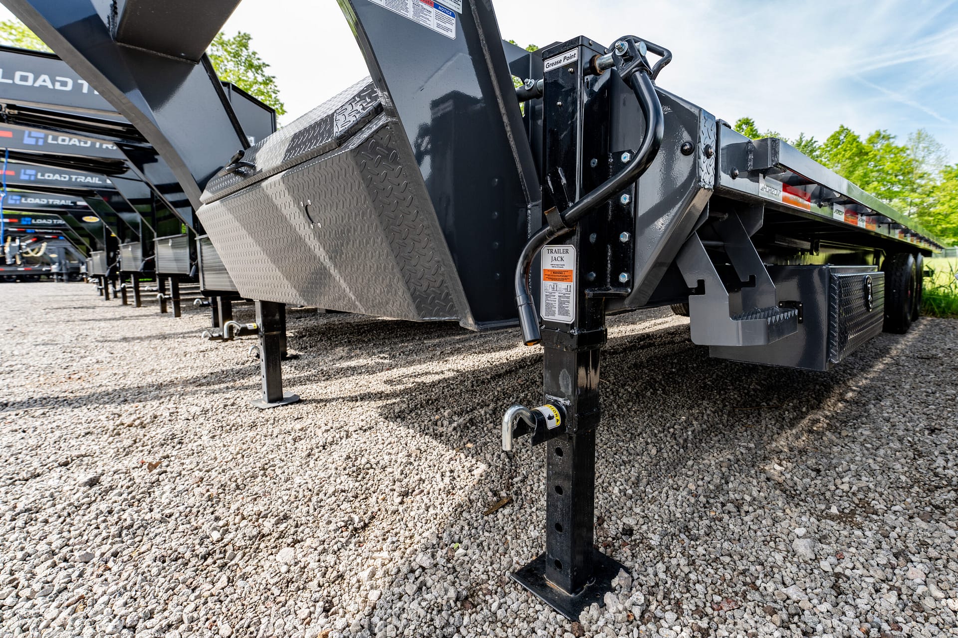 Close-up view of a heavy steel trailer tongue with a raised jack stand, hydraulic hoses, and diamond-plate toolbox on a gravel lot with other trailers in the background.