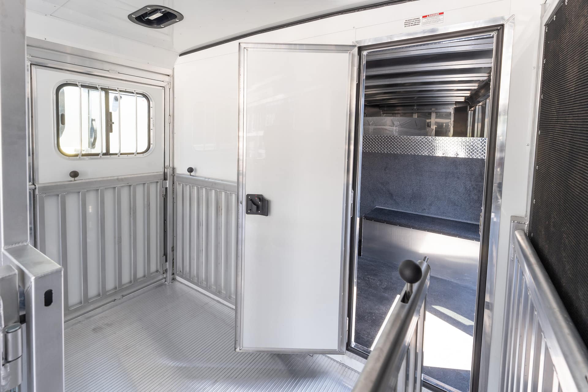 Interior of a silver livestock trailer showing a partition gate, white walls, and a doorway into a cargo area with a diamond-plate step.