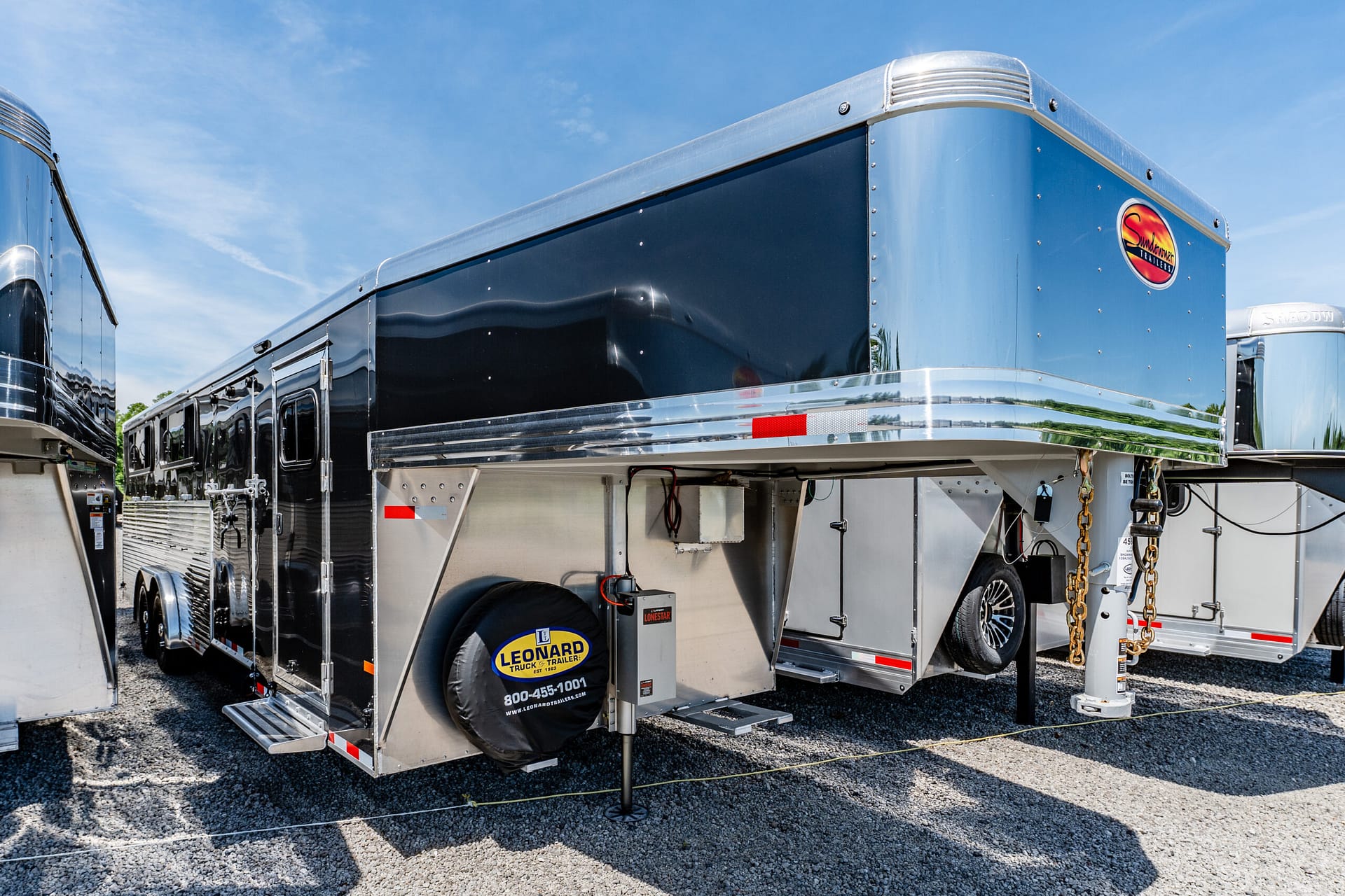 Shiny blue and silver enclosed trailer with chrome trim parked in a lot under a blue sky; logo reads Sundowner Trailers on the side.