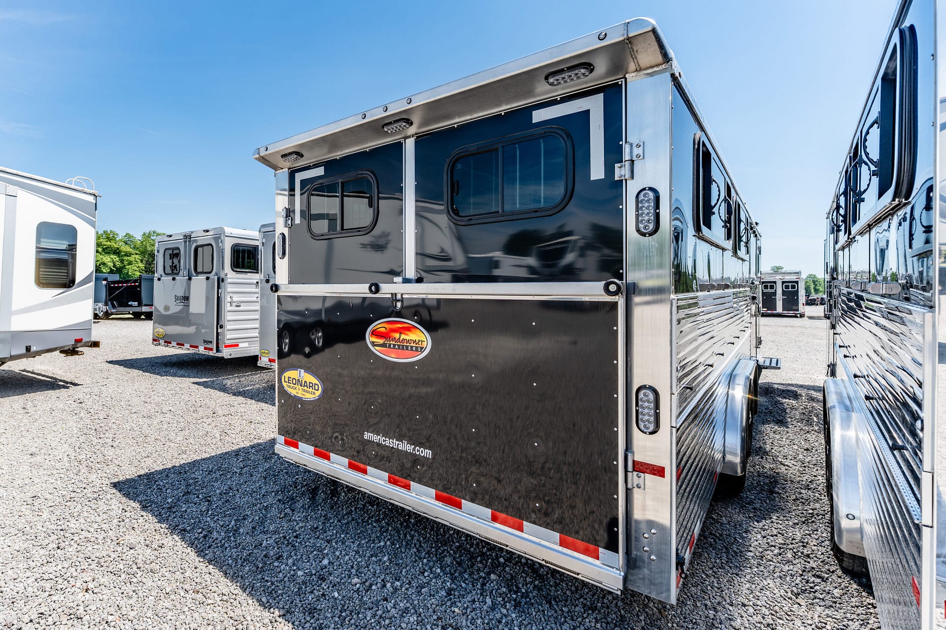 Back of a shiny black Sundowner trailer with chrome trim, parked on gravel among other trailers under a blue sky.