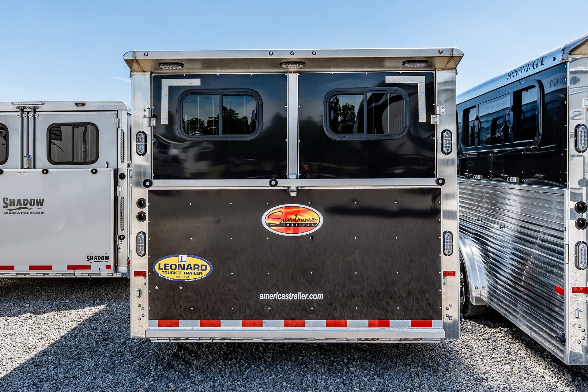 Rear of a shiny black Sundowner trailer with two small windows, metal trim, and company logos visible on a gravel lot.