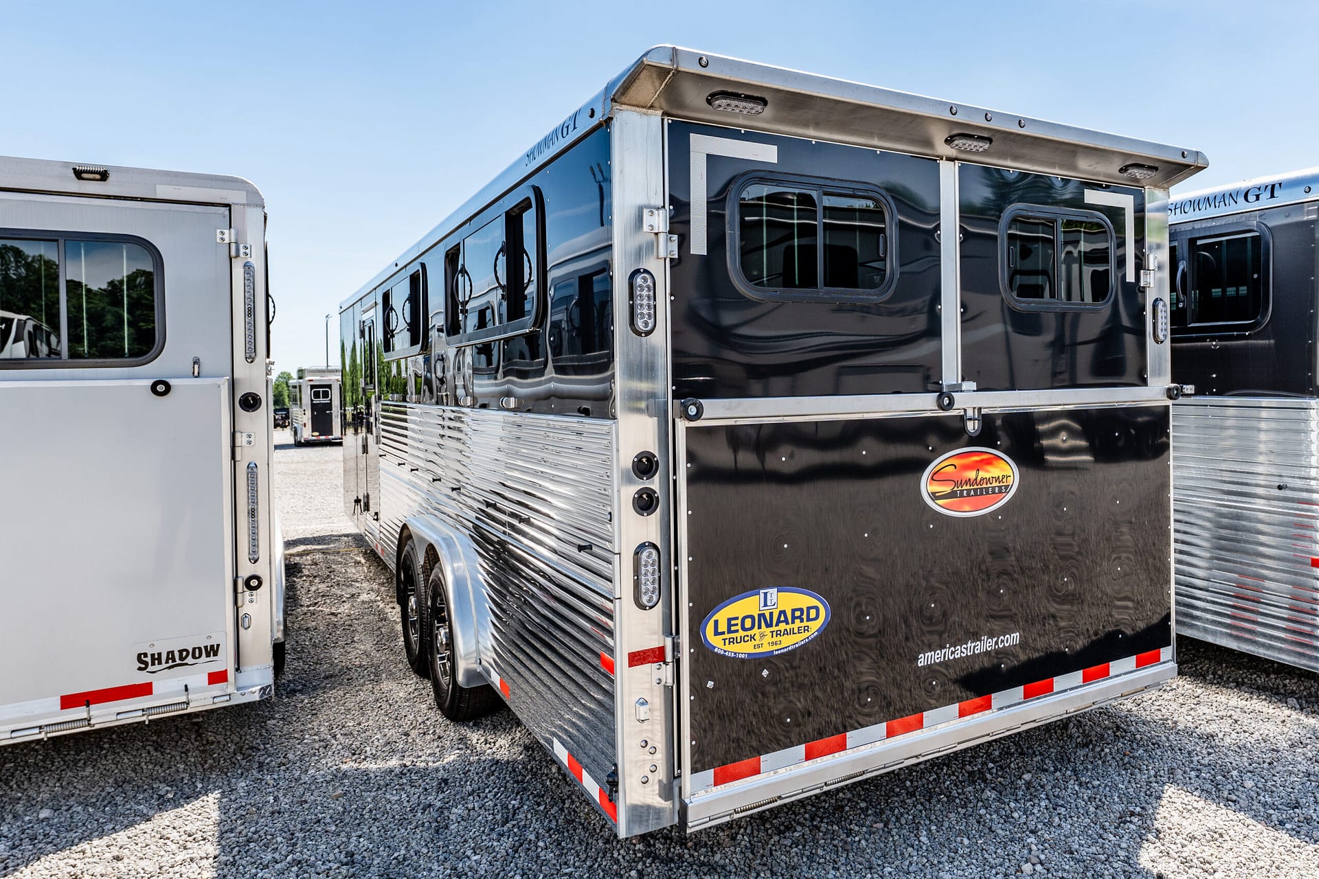 Shiny black aluminum trailer with chrome trim and brand stickers parked on a gravel lot under a blue sky.