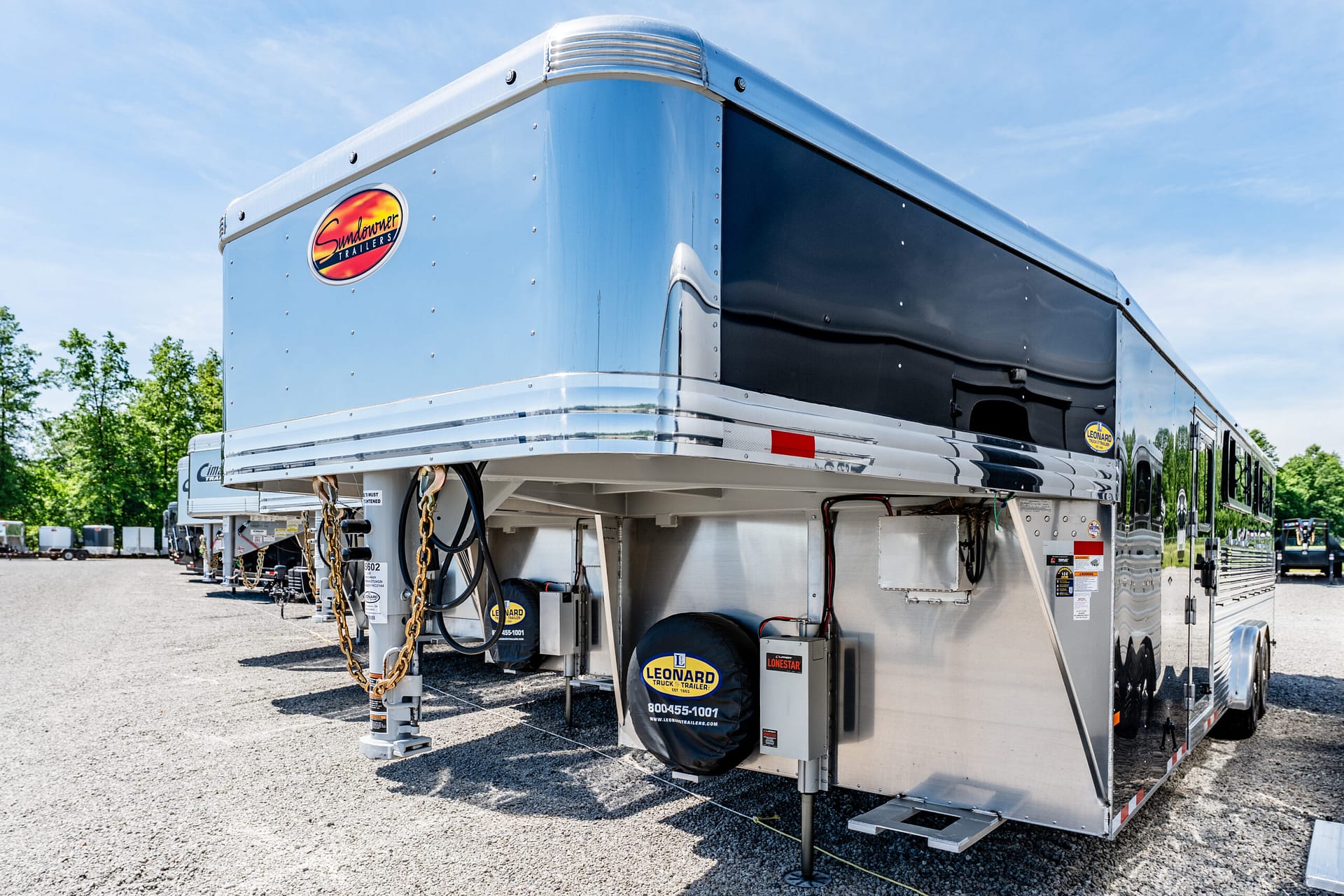 Silver semi-trailer with chrome trim and a Sundowner Trailers logo, parked on a gravel lot under a blue sky.
