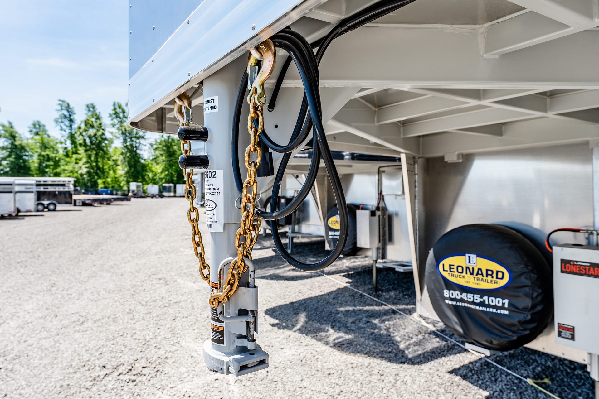 Close-up of a trailer hitch area with gold chains and black cables on a gray trailer, outdoors in a parking lot.