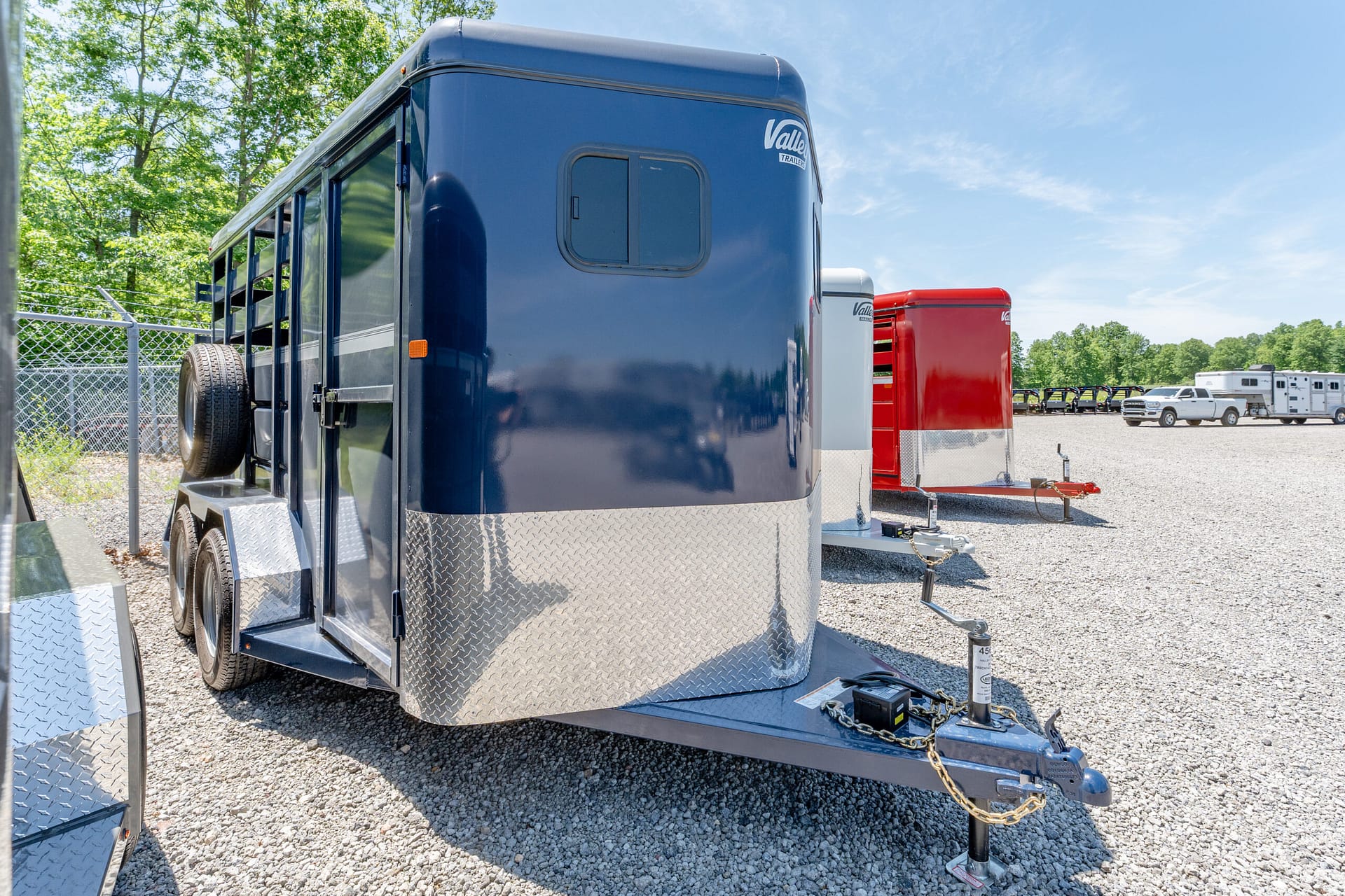 Blue horse trailer with diamond-plate front resting on a gravel lot, with other trailers and a white pickup in the background.