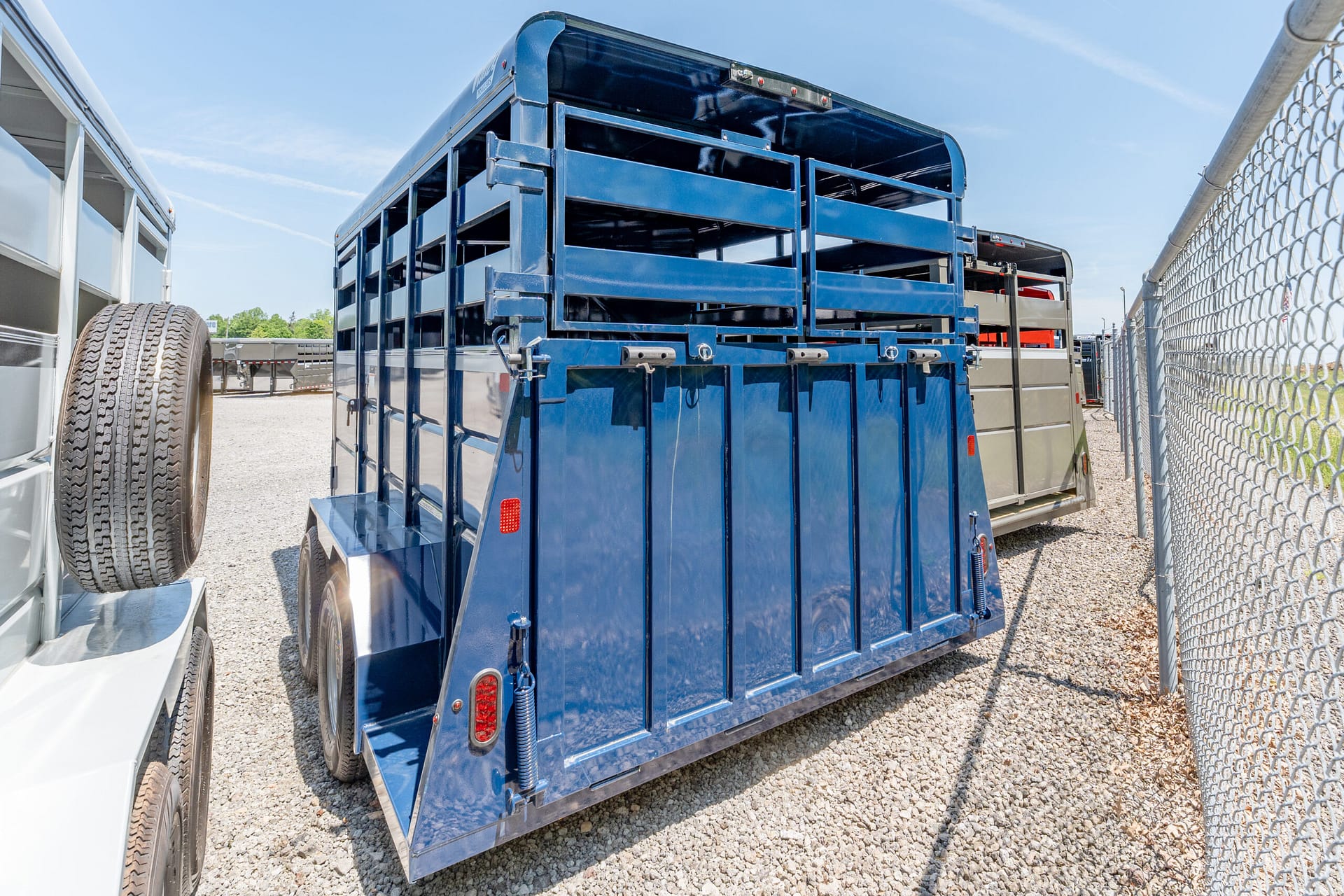 Blue livestock trailer in a gravel lot, with a chain-link fence on the right and other trailers nearby.