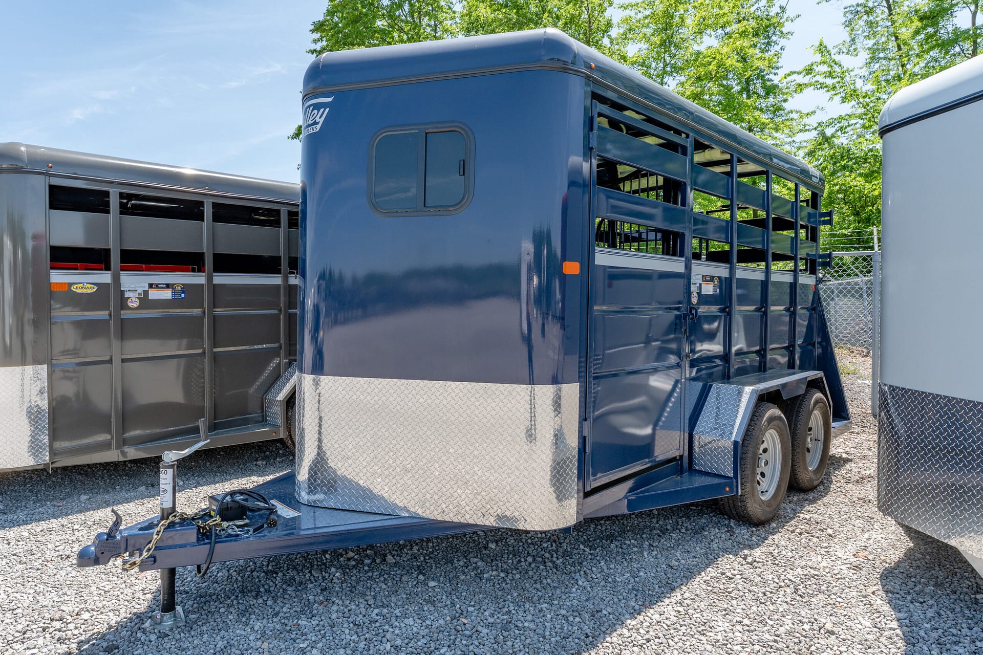 Blue livestock trailer with diamond-plate front, hitched to a tow hitch on gravel, showing tied chains and wheels in foreground.