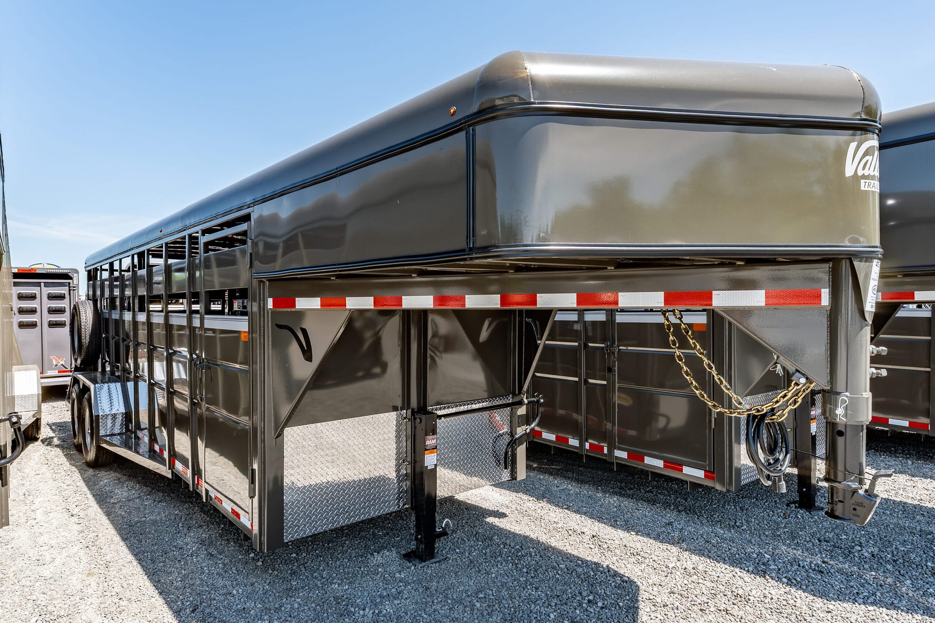 Large gray livestock trailer with metal body, open side panels, red/white reflective tape, and safety chains on a gravel lot.