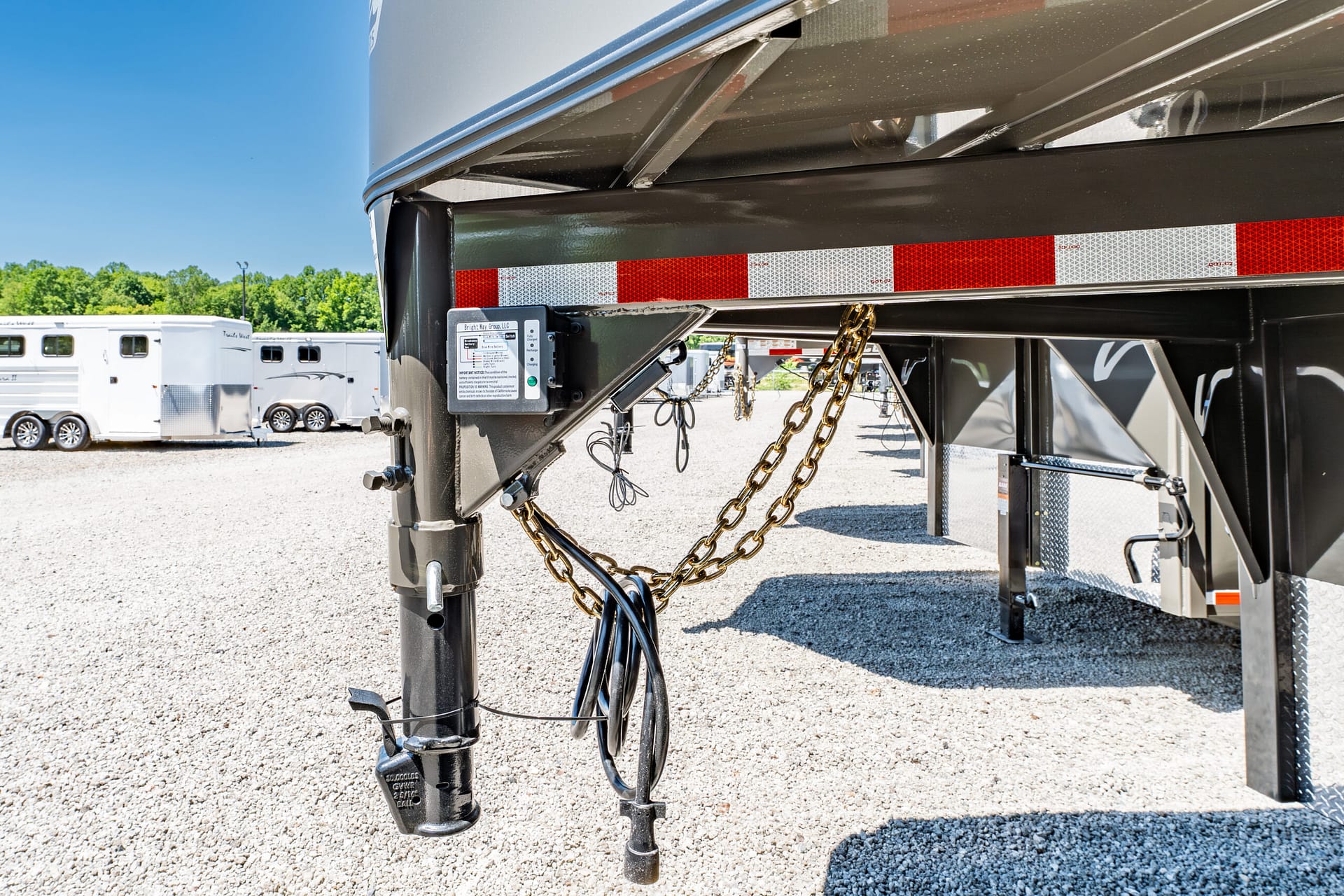 Close-up of a gooseneck trailer hitch with safety chains and hydraulic lines hanging below, on a gravel lot with other trailers in the background
