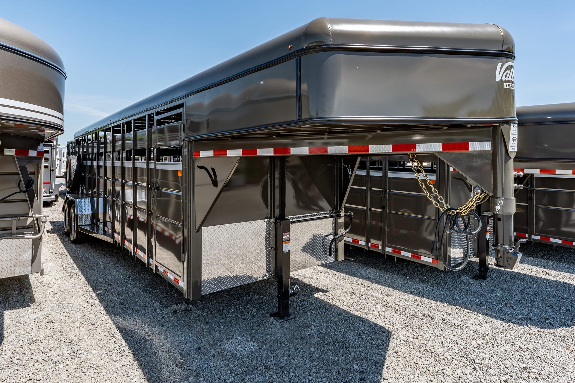 Black horse trailer with open side ventilation vents, reflective stripes, and diamond-plate entry steps, parked on gravel under a clear blue sky.