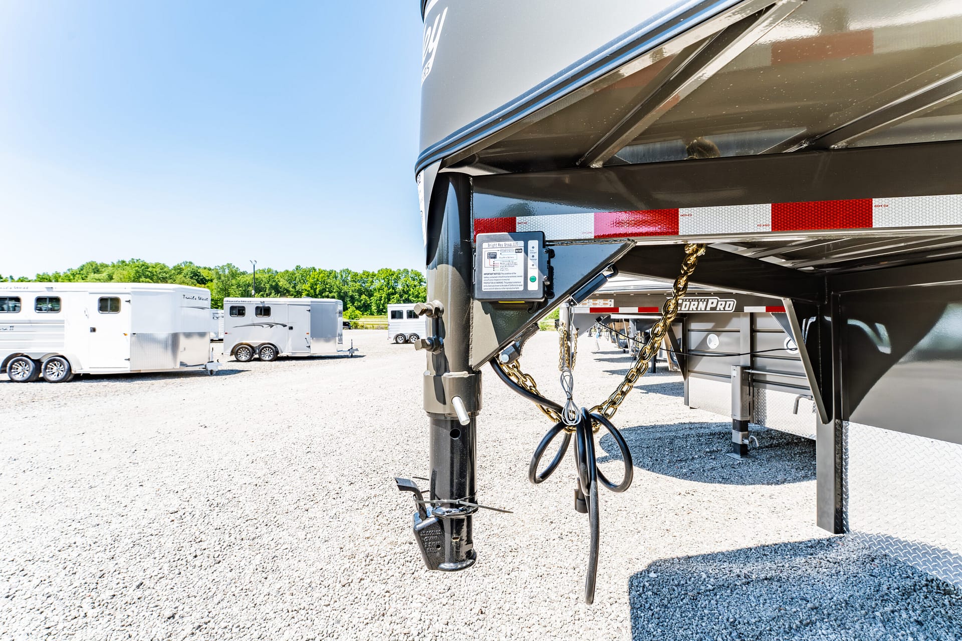 Close-up of a gooseneck trailer hitch with safety chains and hydraulic jack, parked in a gravel lot with other horse trailers in the background.