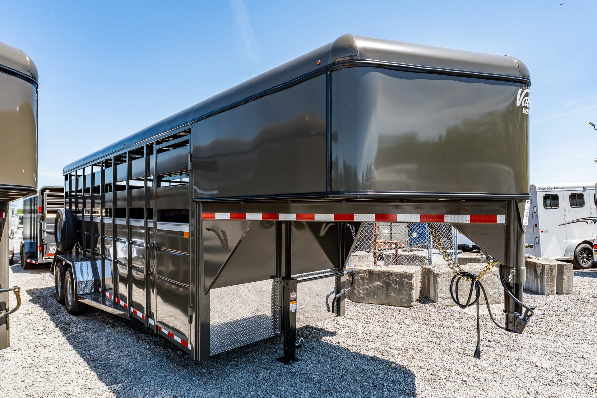 Black gooseneck livestock trailer parked on a gravel lot with visible wheels and chrome diamond-plate steps at the rear (outdoor daylight).