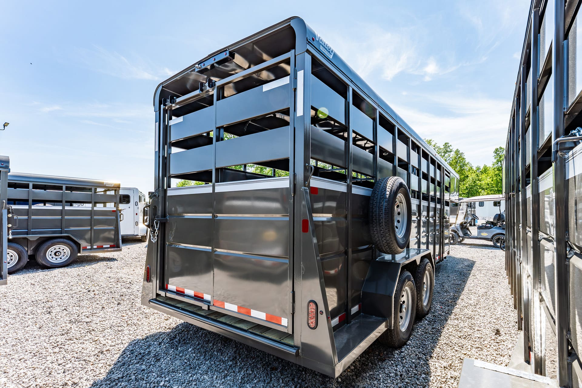 Large gray livestock trailer with open side slats and a spare tire mounted on the side, parked among other trailers on a gravel lot under a blue sky.