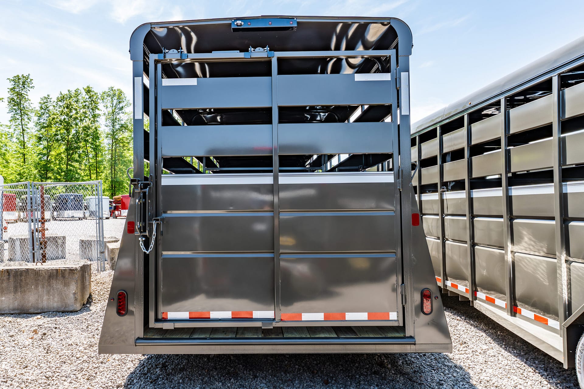 Rear view of a gray metal livestock trailer with its back doors open, parked on gravel yard under a clear sky.