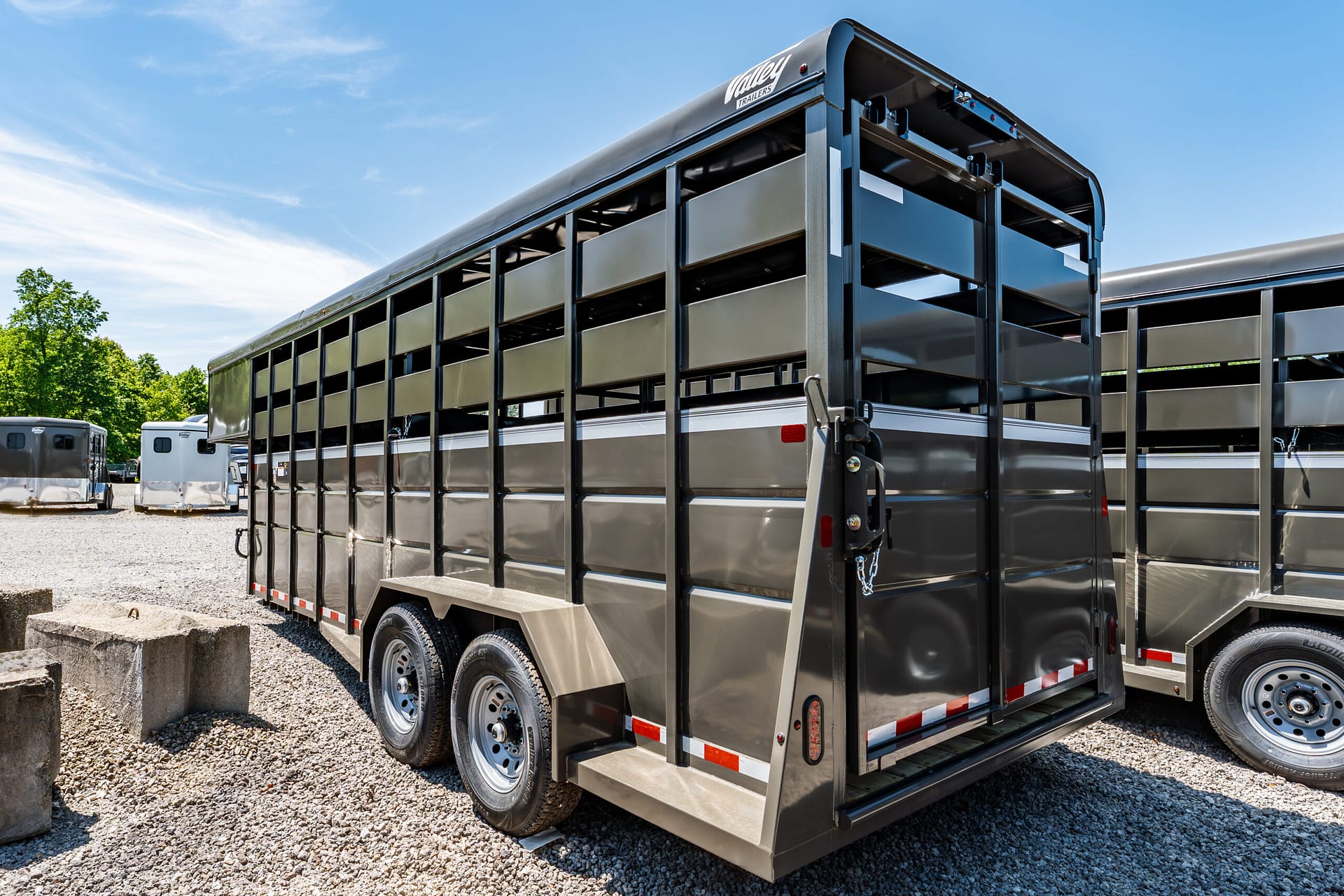 Side view of a large black metal livestock trailer parked on a gravel lot under a bright blue sky.