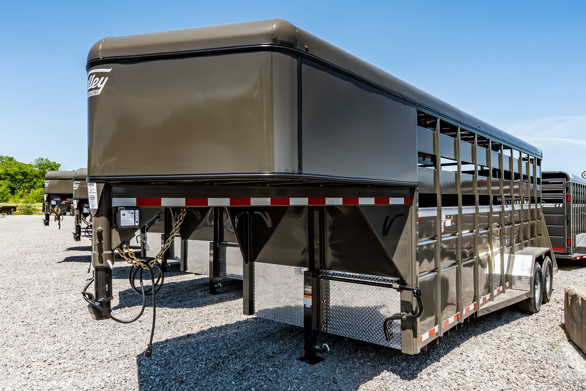Large dark gray gooseneck trailer on a gravel lot, with open side windows and red-and-white reflective stripes along the bottom edge, under a clear blue sky