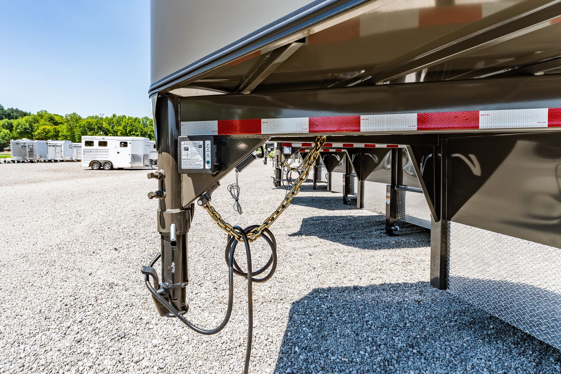 Close-up of a trailer hitch with safety chains and electrical cables under a black trailer, on a gravel lot with other trailers in the background.