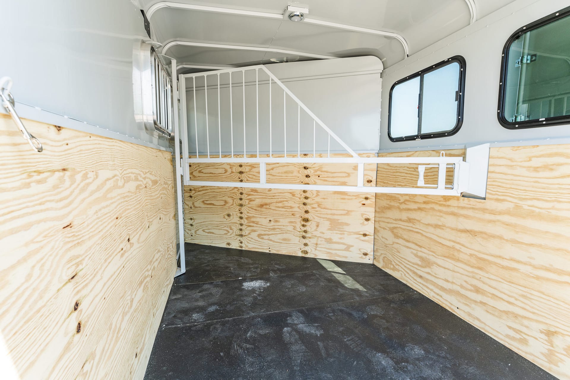 Interior of an empty horse trailer with light wood walls, a white metal divider, and a small window on the right side of the stall area.