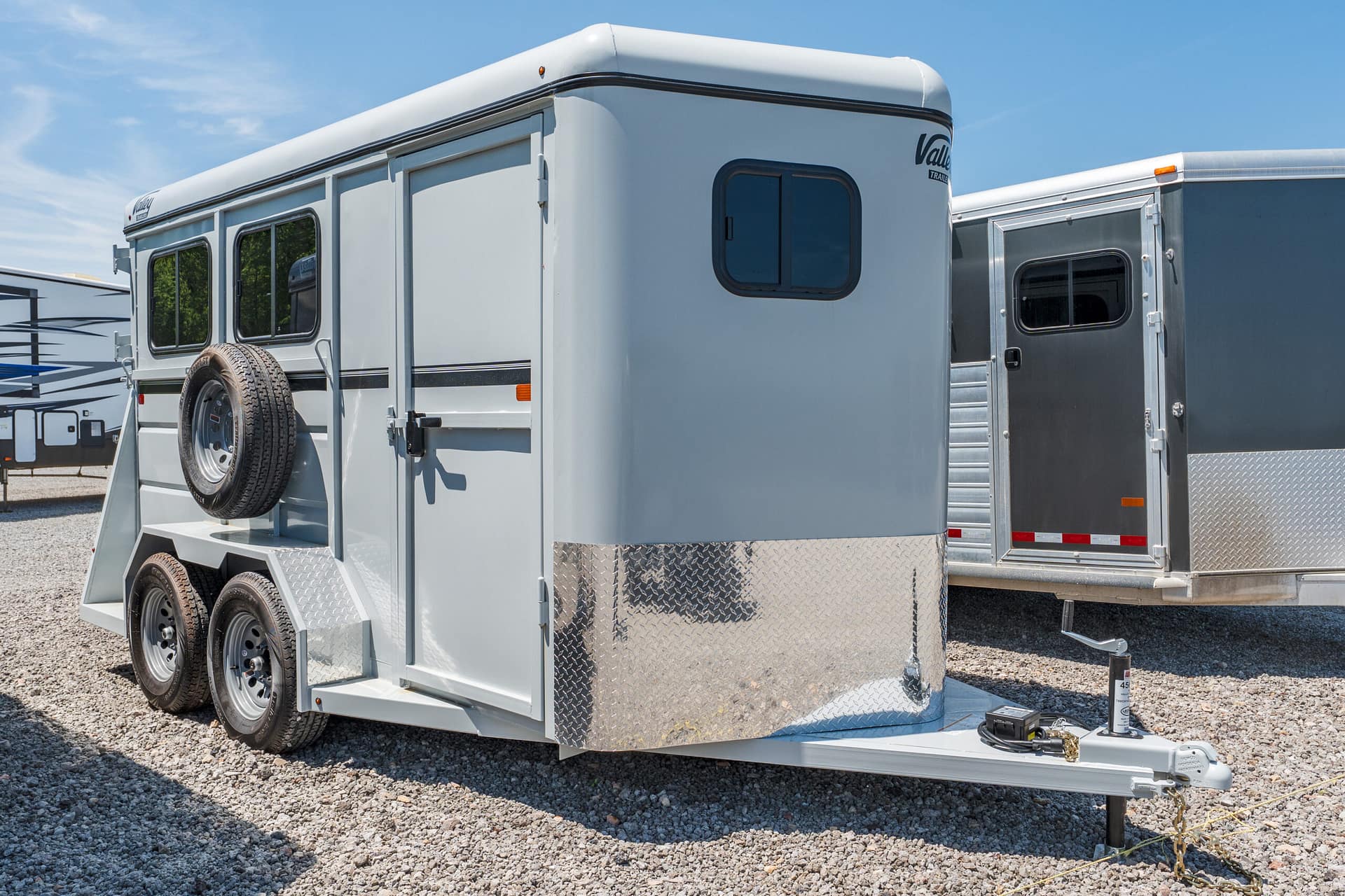 Silver horse trailer with dual axles and a mounted spare tire on the side, parked on gravel under a clear blue sky