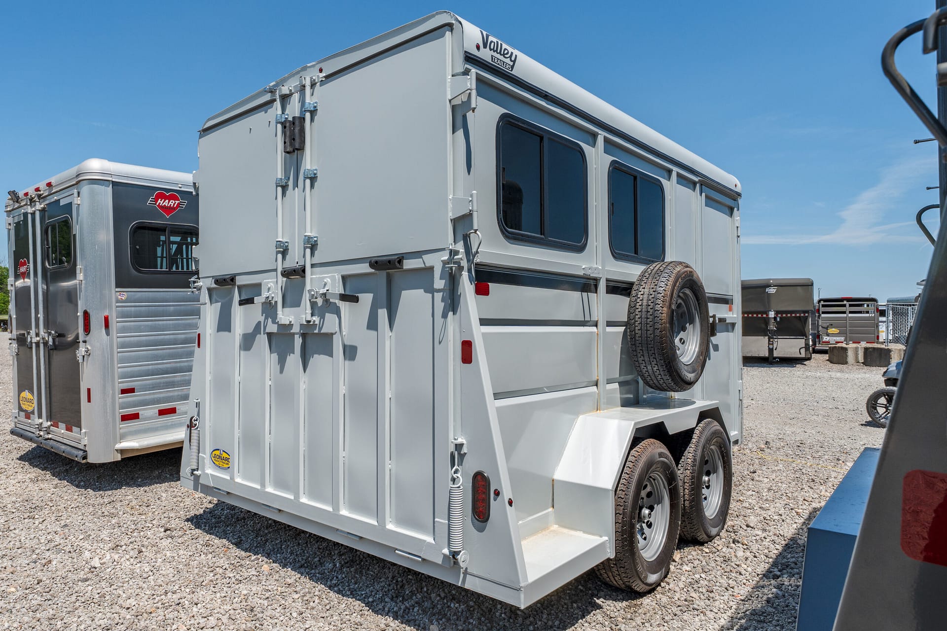 Grey livestock/horse trailer with two side windows and a spare tire mounted on the back, parked on a gravel lot under a clear blue sky.