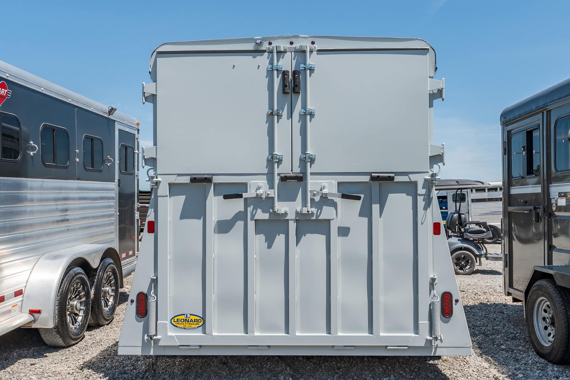 Rear view of a large gray cargo trailer with double doors and heavy latches, flanked by other trailers on a gravel lot under a clear blue sky.