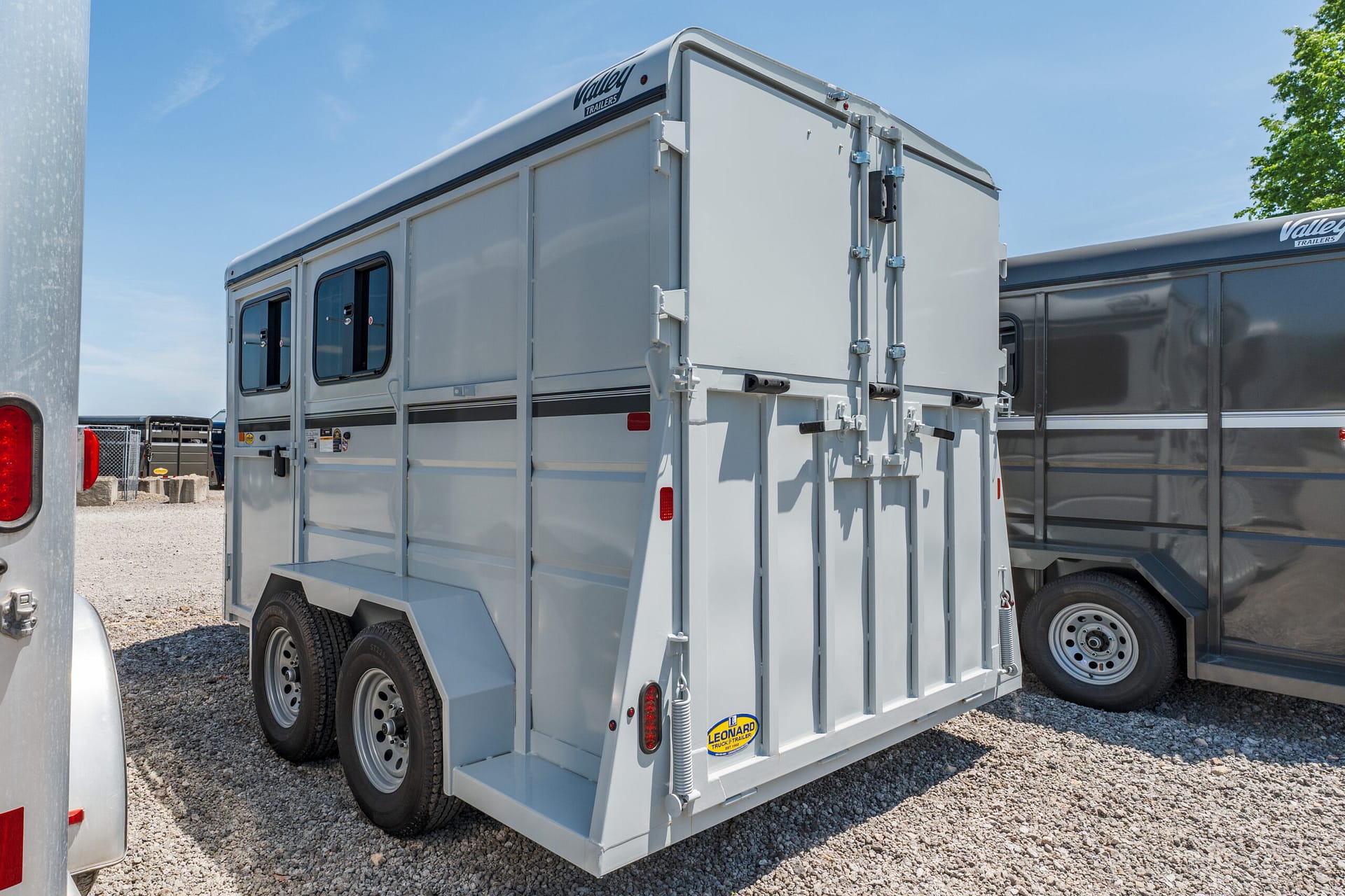 Rear view of a light gray livestock trailer with dual doors and latching hardware, parked on a gravel lot under a blue sky.