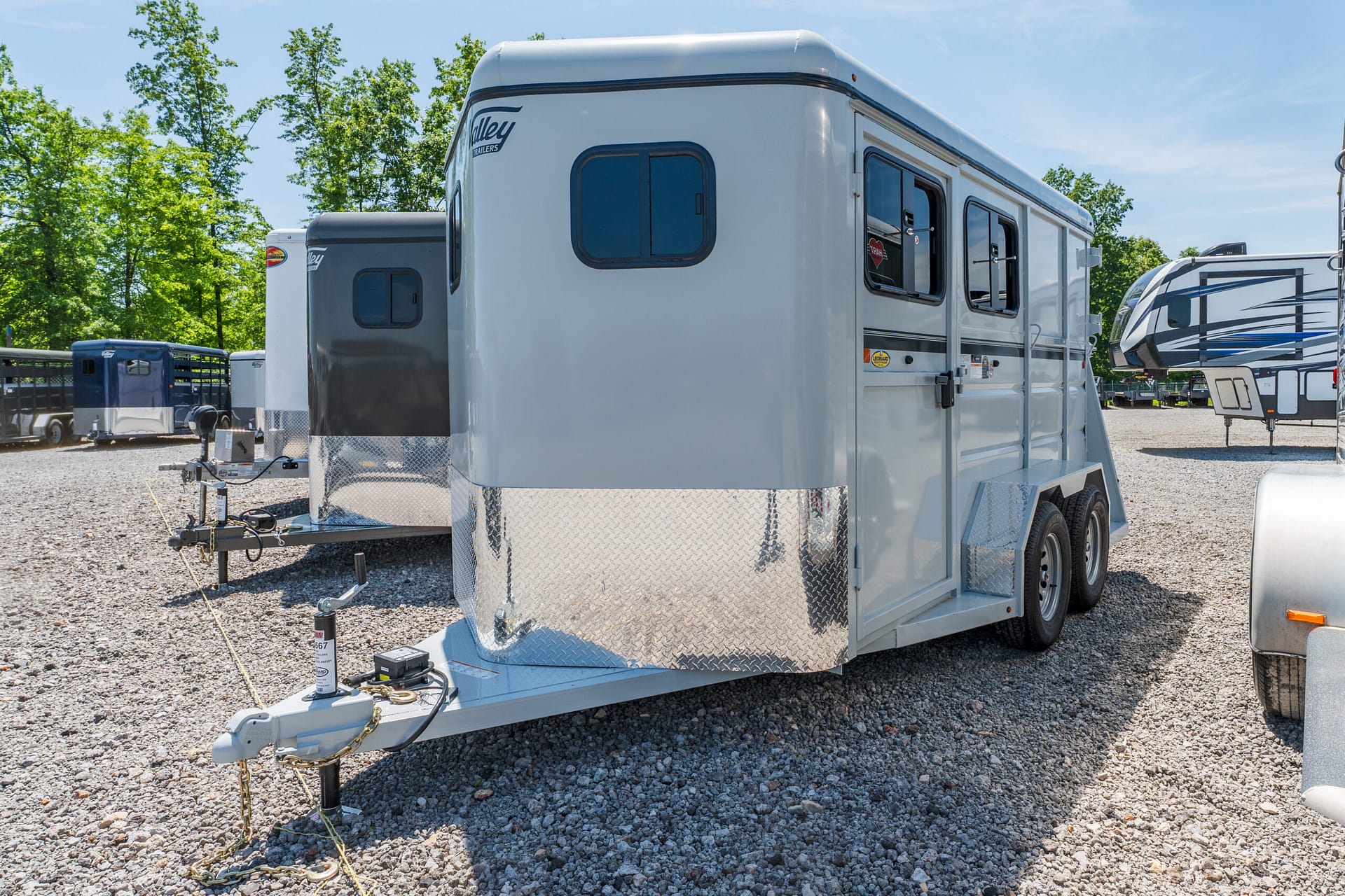 Silver travel trailer with a front hitch on a gravel lot, trees in the background.
