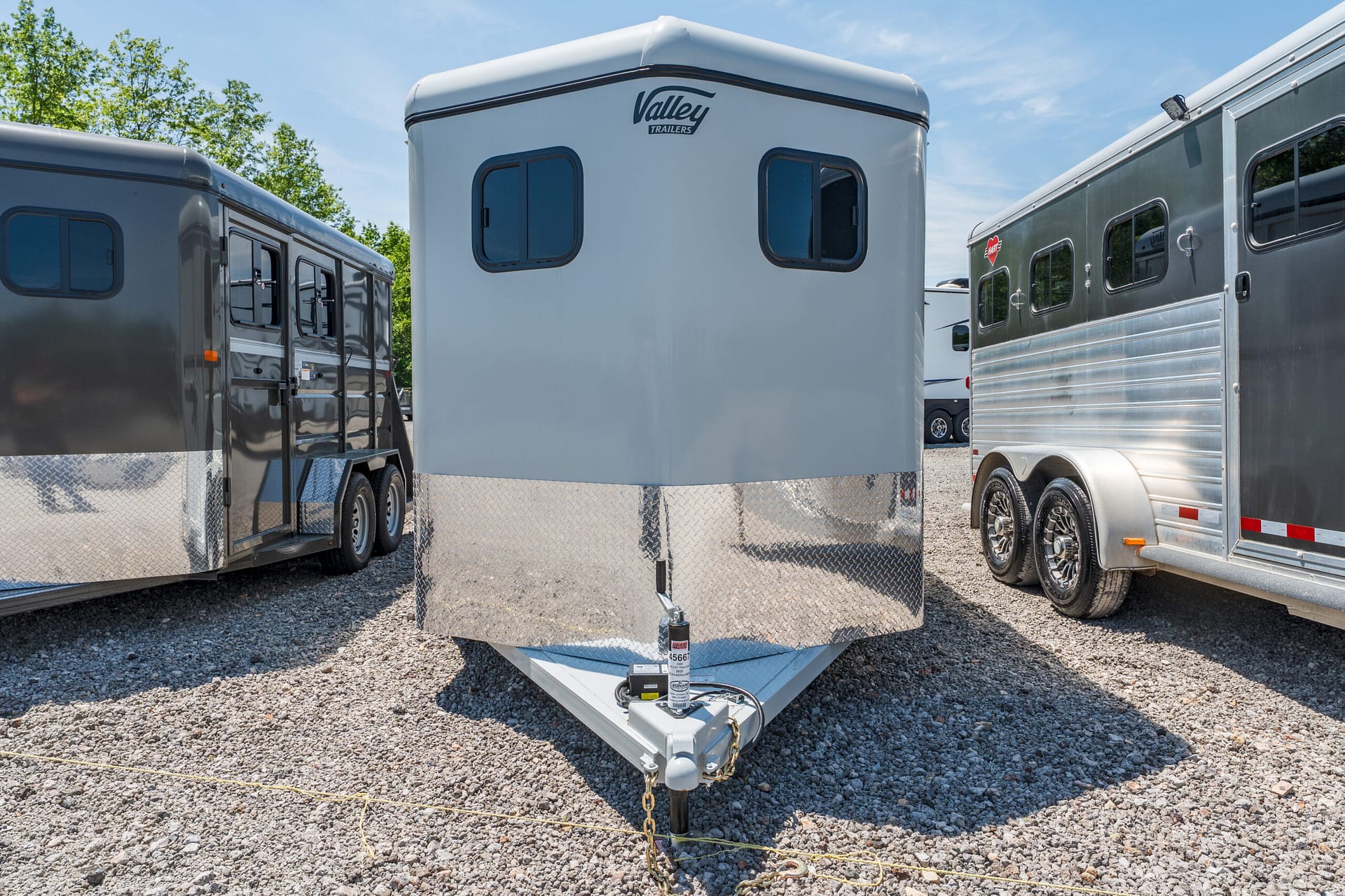 White horse trailer centered between two other trailers on a gravel lot under a blue sky, with a Valley Trailers logo visible on the front.
