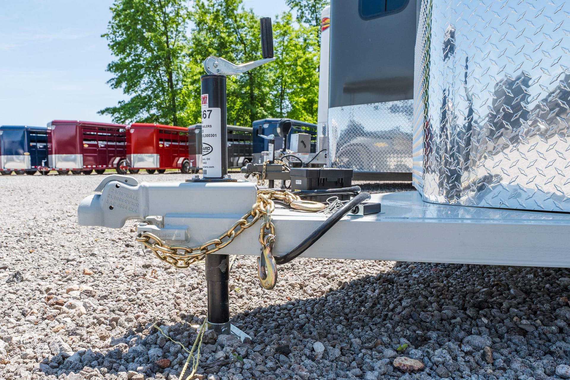 Close-up of a trailer hitch with safety chains and a handle, set on a gravel lot with colorful trailers in the background.