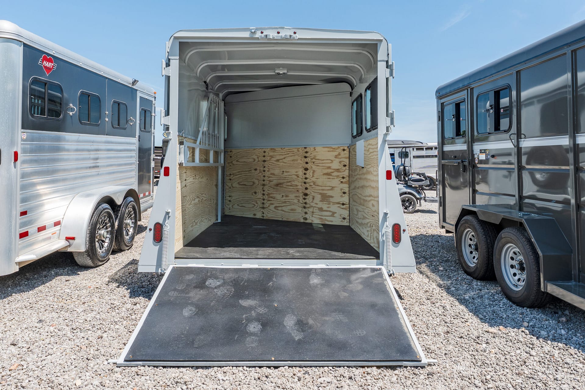 Open horse trailer with ramp down, showing wooden walls and partition, parked between other trailers on gravel.