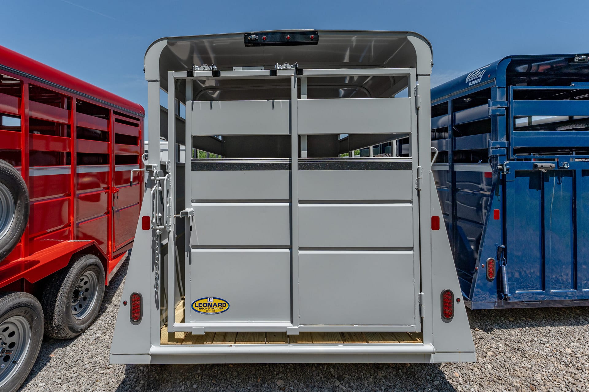 Rear of a gray livestock trailer with its doors open, showing interior stalls, between red and blue trailers on a gravel lot under a clear blue sky (Leonard logo on the bumper).