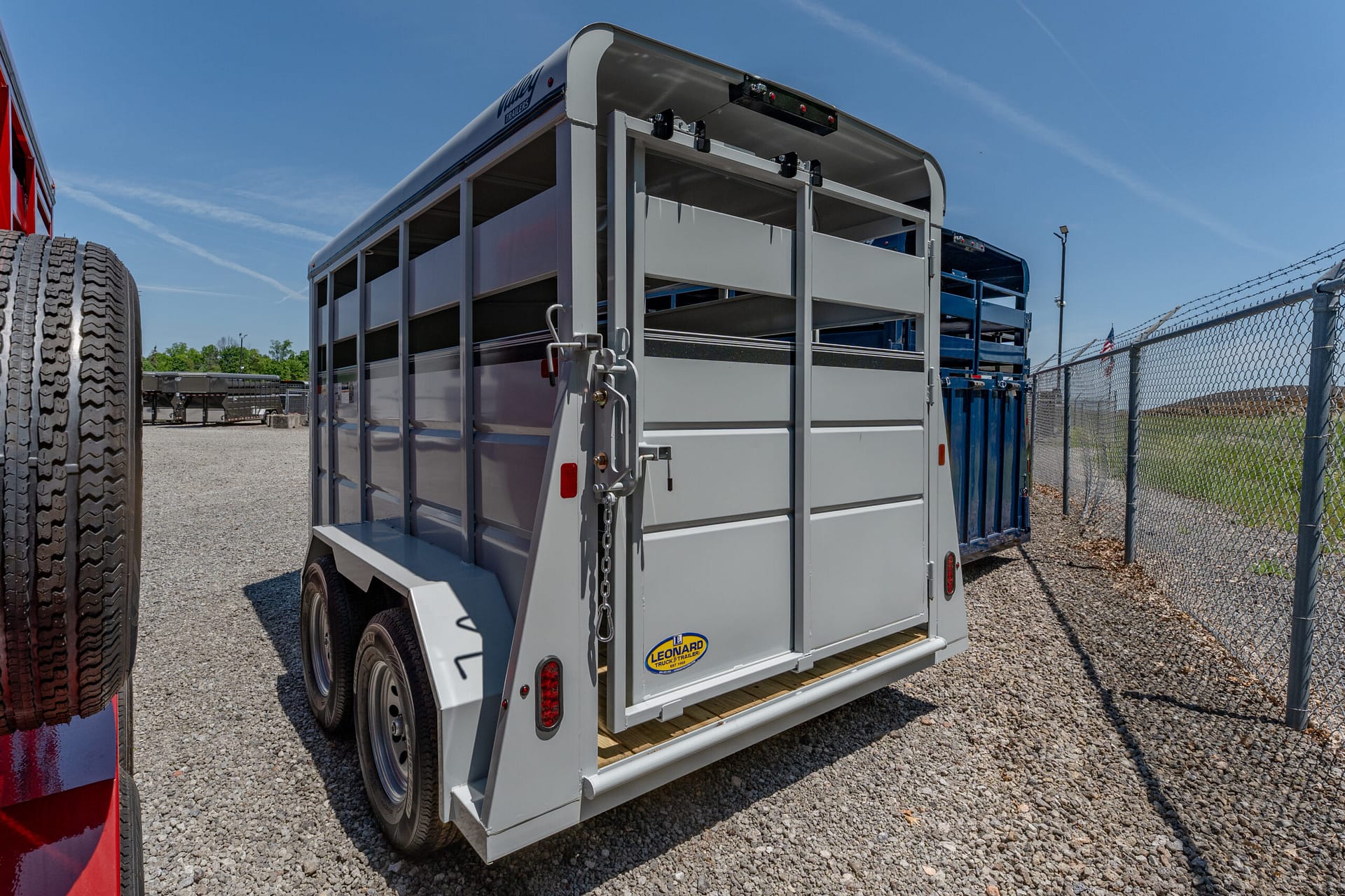 Rear view of a silver livestock trailer with open side panels parked on a gravel lot under a blue sky.