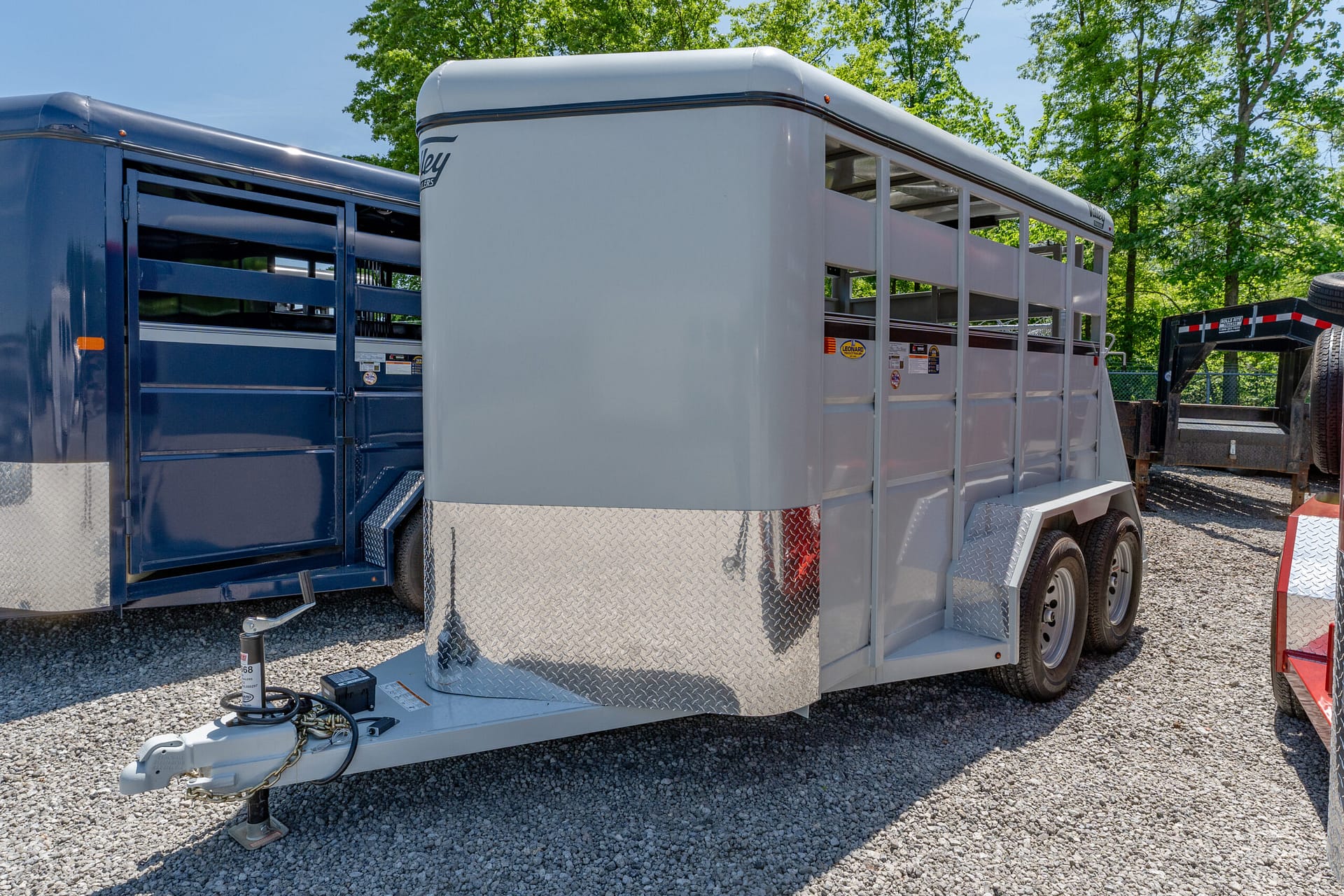 Light gray livestock trailer with diamond-plate front on a gooseneck hitch, parked on gravel with a blue trailer beside it and green trees in the background.