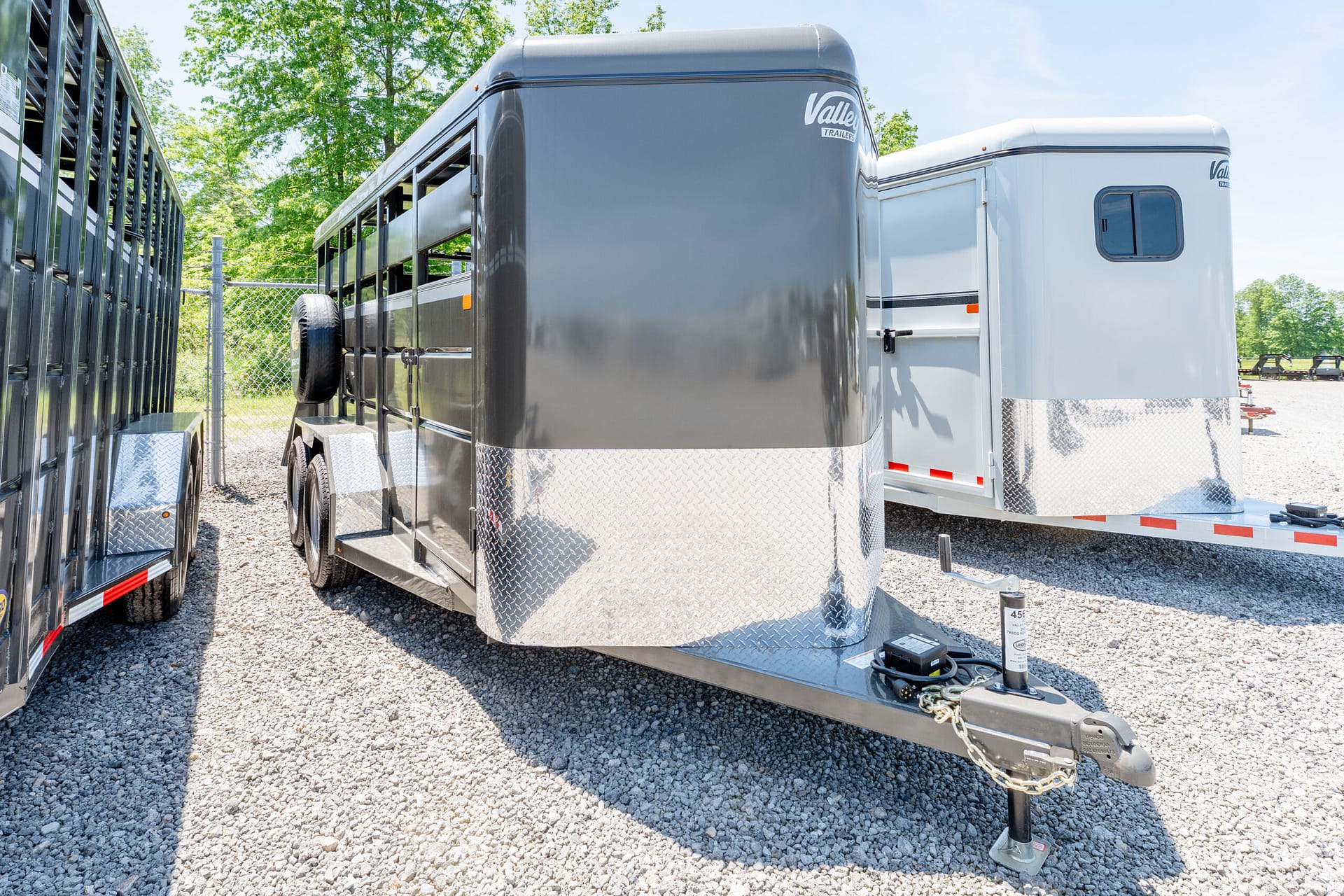 A gray travel trailer with diamond-plate front on a gravel lot, hitch and safety chains visible in the foreground with a fence nearby.