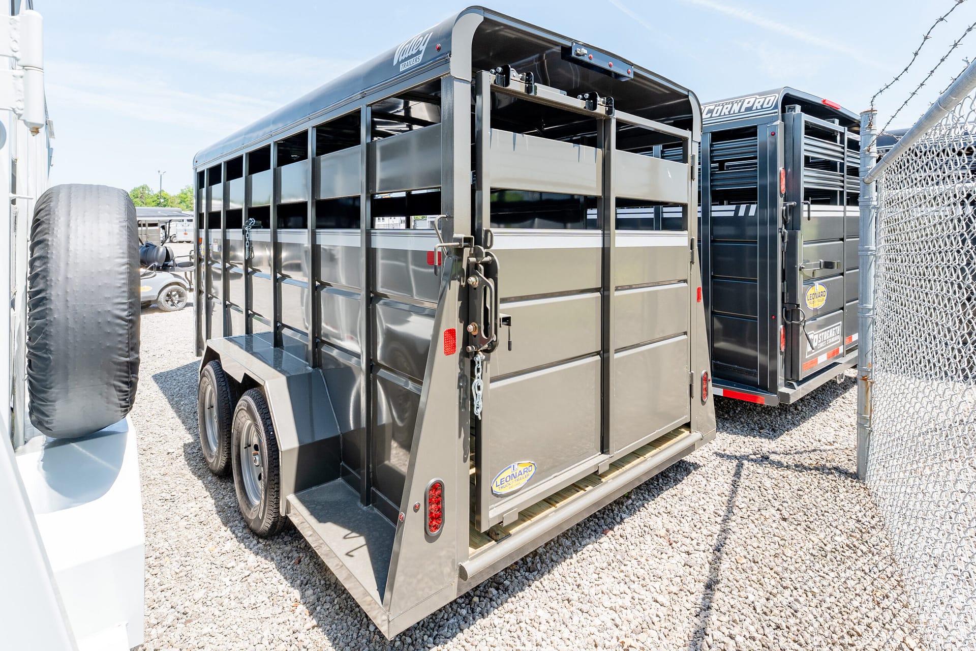Side view of a gray livestock trailer with open side gates in a gravel lot, spare tire nearby, and barbed fence in background