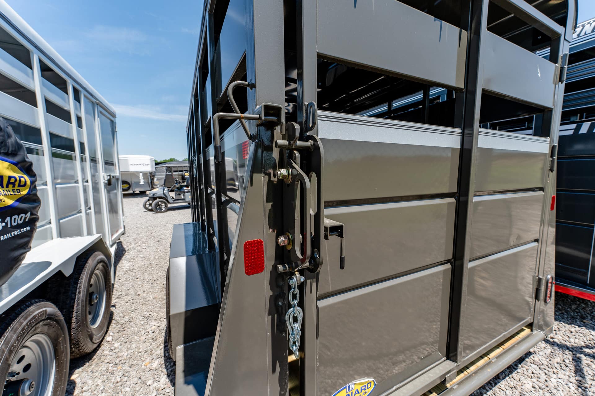 Close-up of a gray metal trailer door with a chain latch and red reflector, other trailers visible in a gravel lot behind.