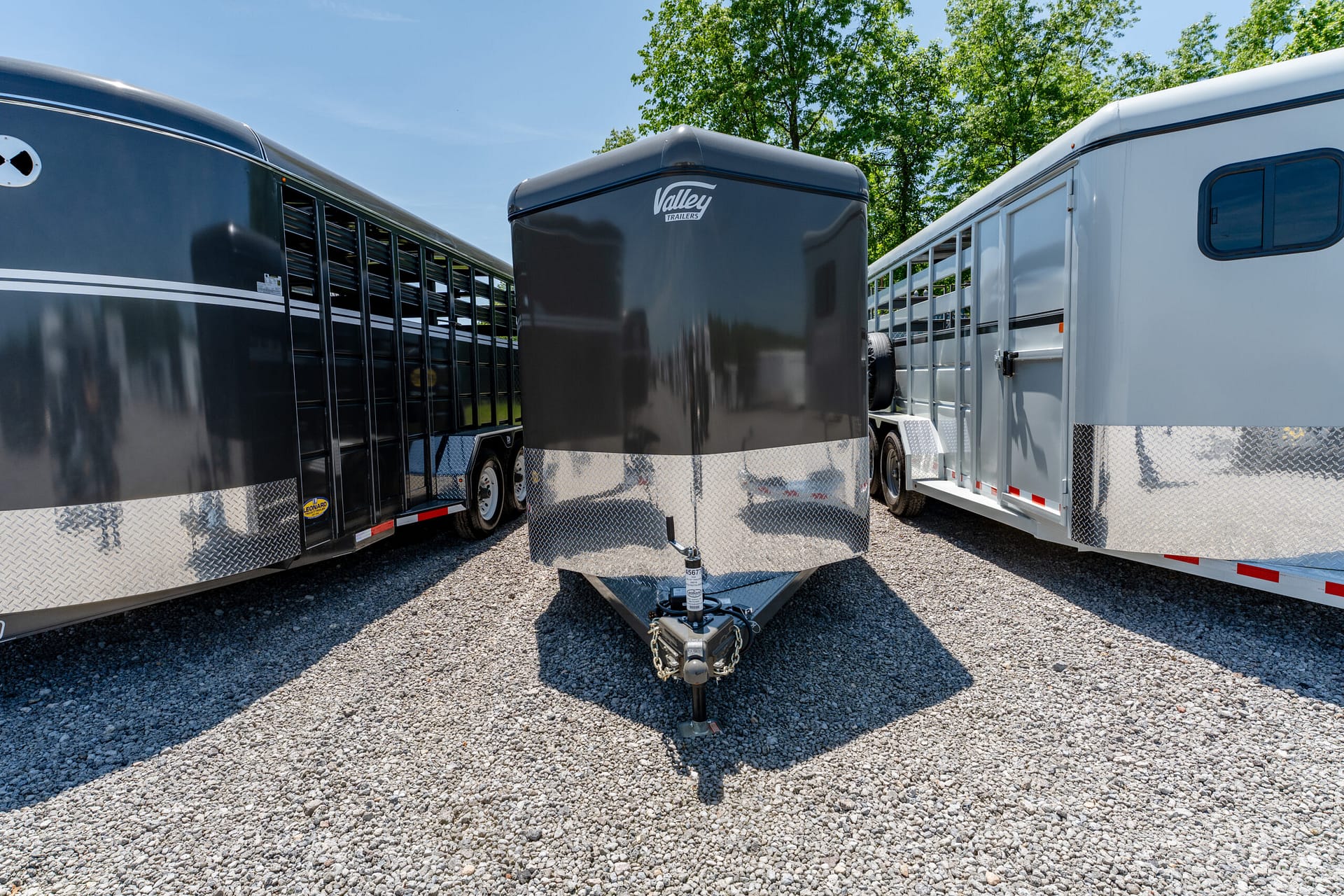Three horse trailers parked on a gravel lot, with a gray Valley trailers unit in the center and a white trailer to the right, blue sky and trees in the background.