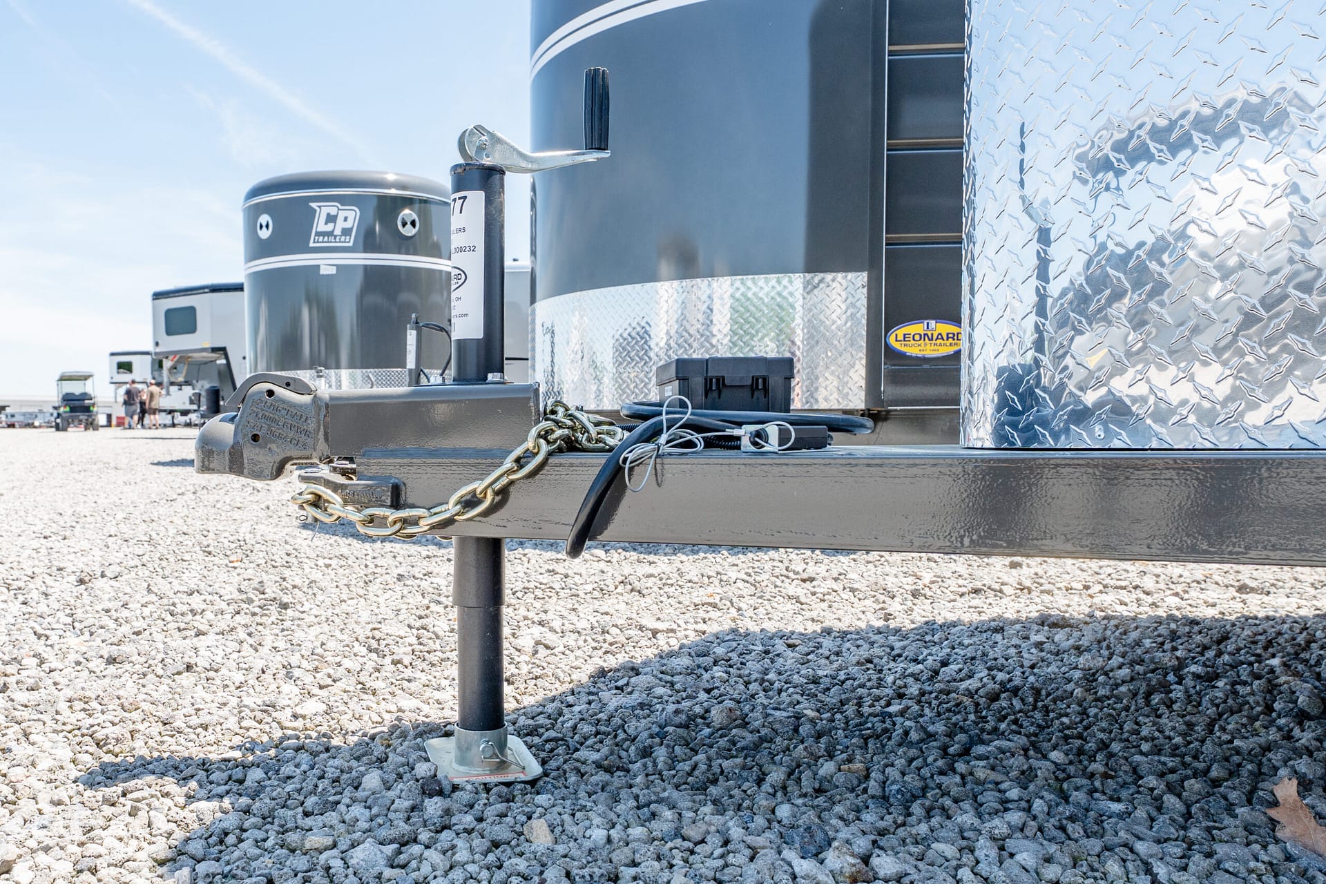 Close-up of a trailer hitch with safety chains and a metal stand on a gravel lot, with diamond-plate trailer siding in the background