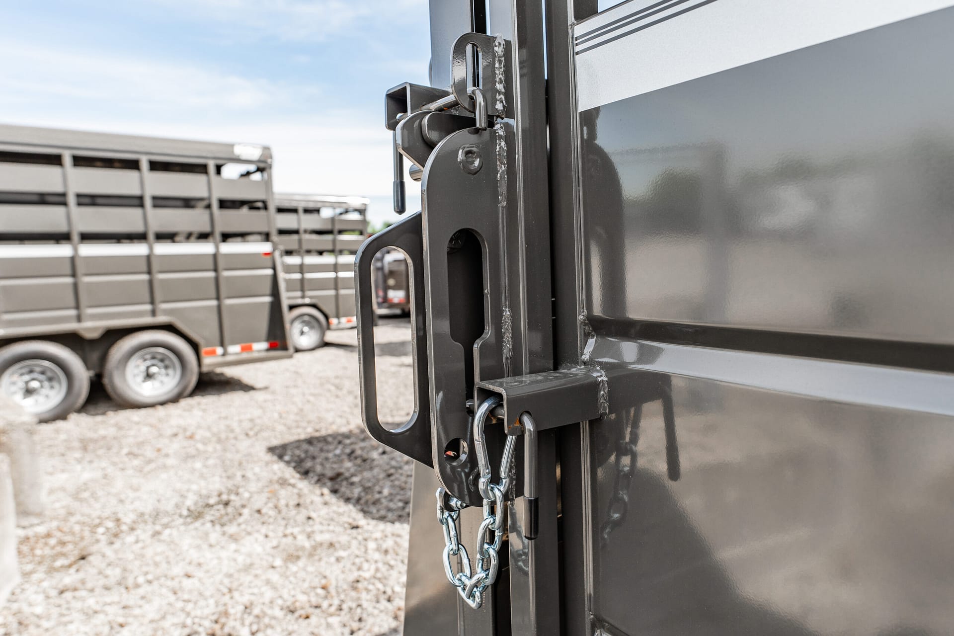 Close-up of a padlock and chain securing a trailer latch on a dark gray trailer, with stacked trailers in a gravel yard in the background.