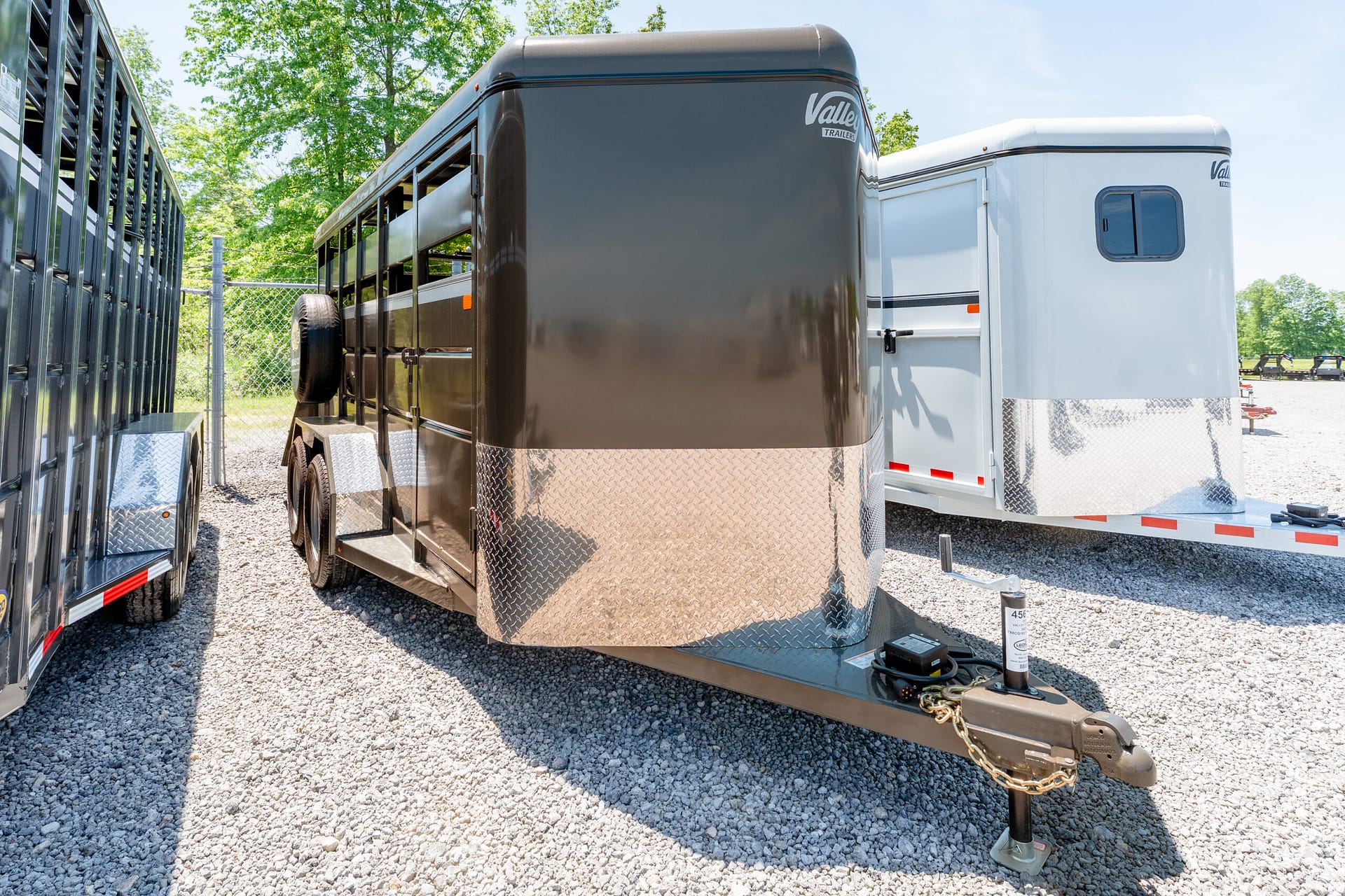 Two large enclosed trailers parked side by side on a gravel lot, with diamond-plate metal fronts and a visible hitch and chains in the foreground.