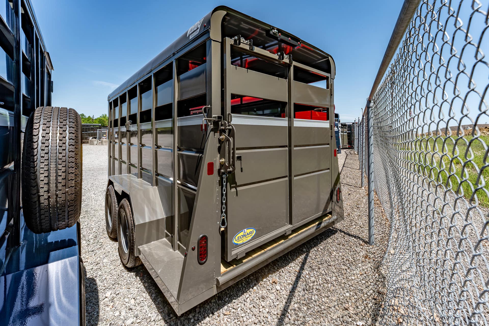 Gray livestock trailer with rear double doors, parked in a gravel lot beside a chain-link fence on a sunny day.