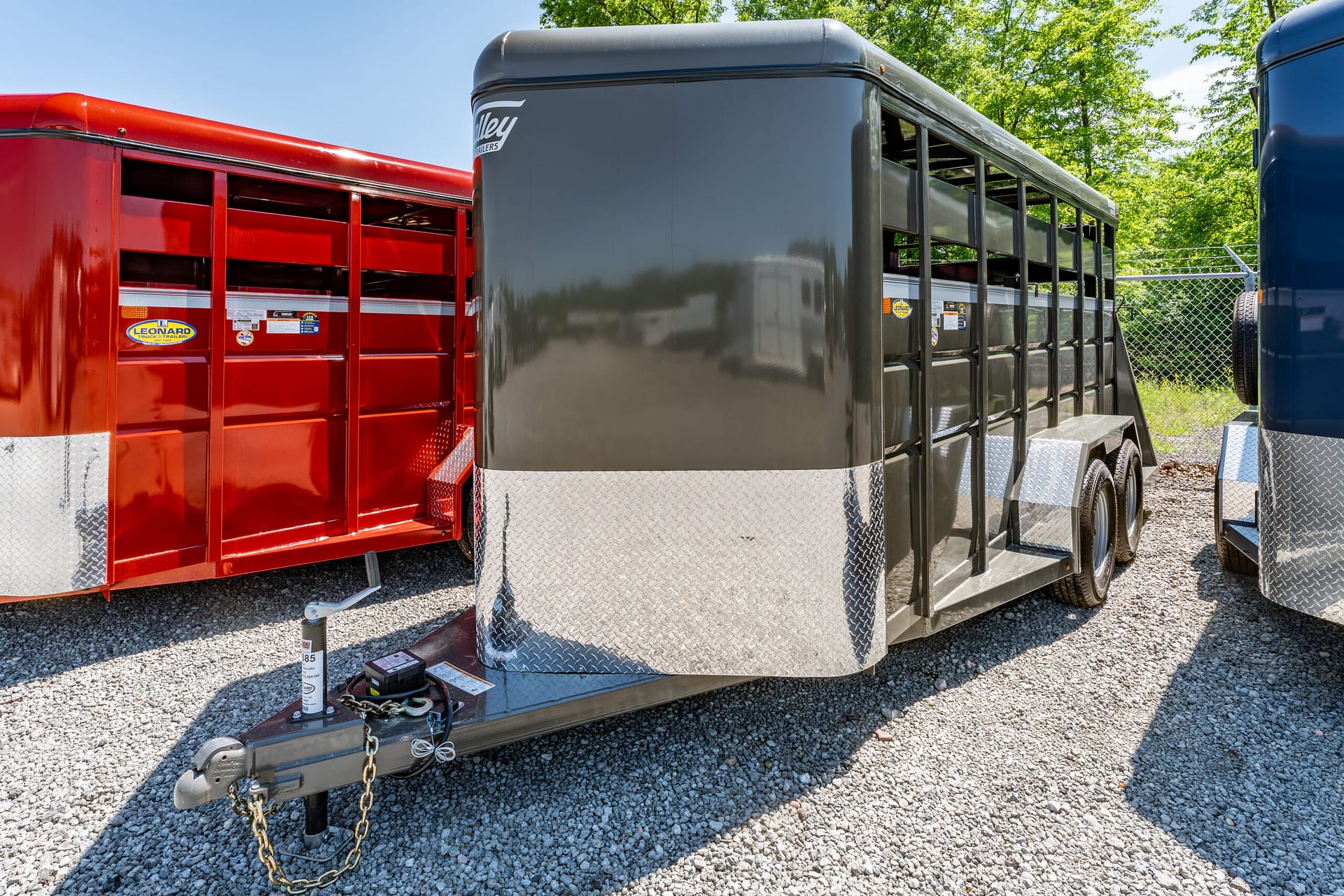 Close-up of a gooseneck trailer hitch on a gravel lot, flanked by a red trailer on the left and a gray trailer on the right.