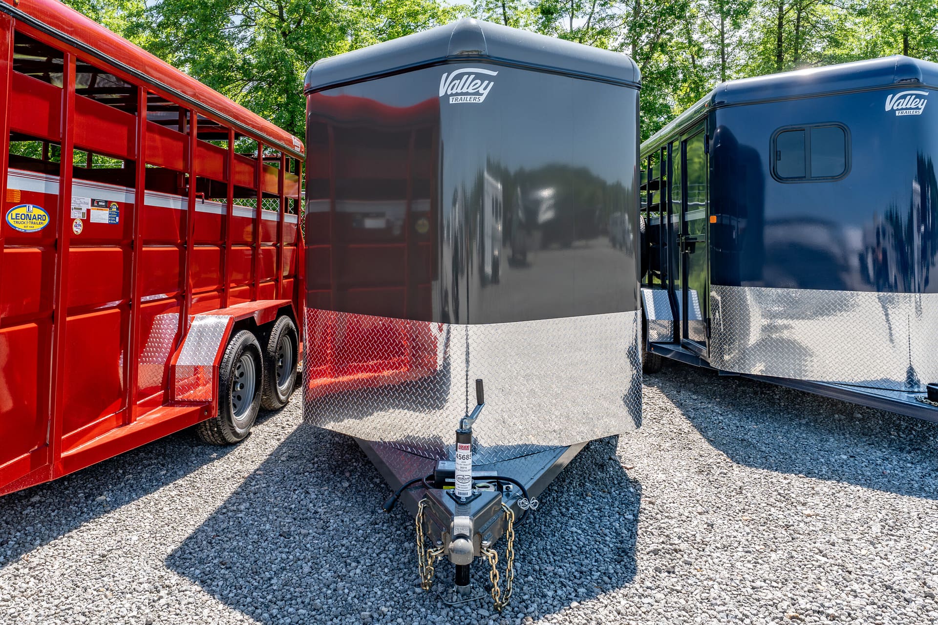 Row of trailers on a gravel lot: red open trailer left, gray and blue enclosed trailers center and right; hitch and chains in foreground.
