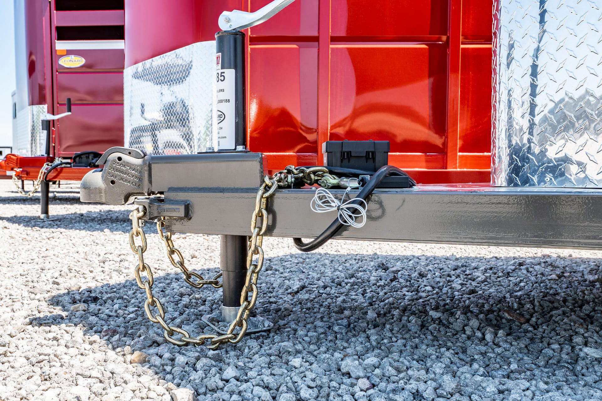 Close-up of a trailer hitch with chains and a jack on a gravel surface, next to a red trailer with diamond-plate metal.