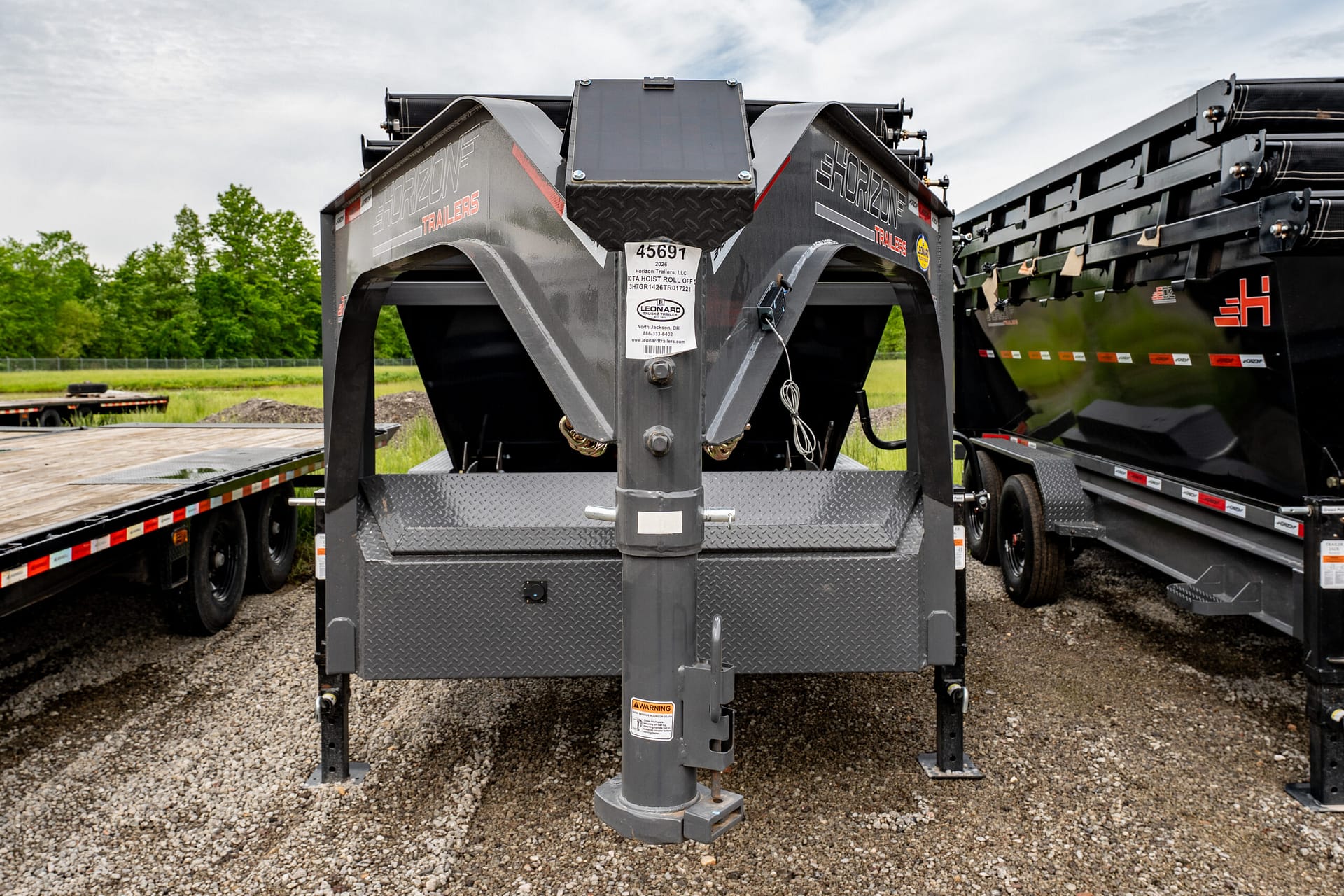Close-up of a heavy-duty gooseneck trailer hitch with diamond-plate steel stairs, warning labels, and a central vertical post, set in a gravel lot with a black trailer on the right and green trees in the background