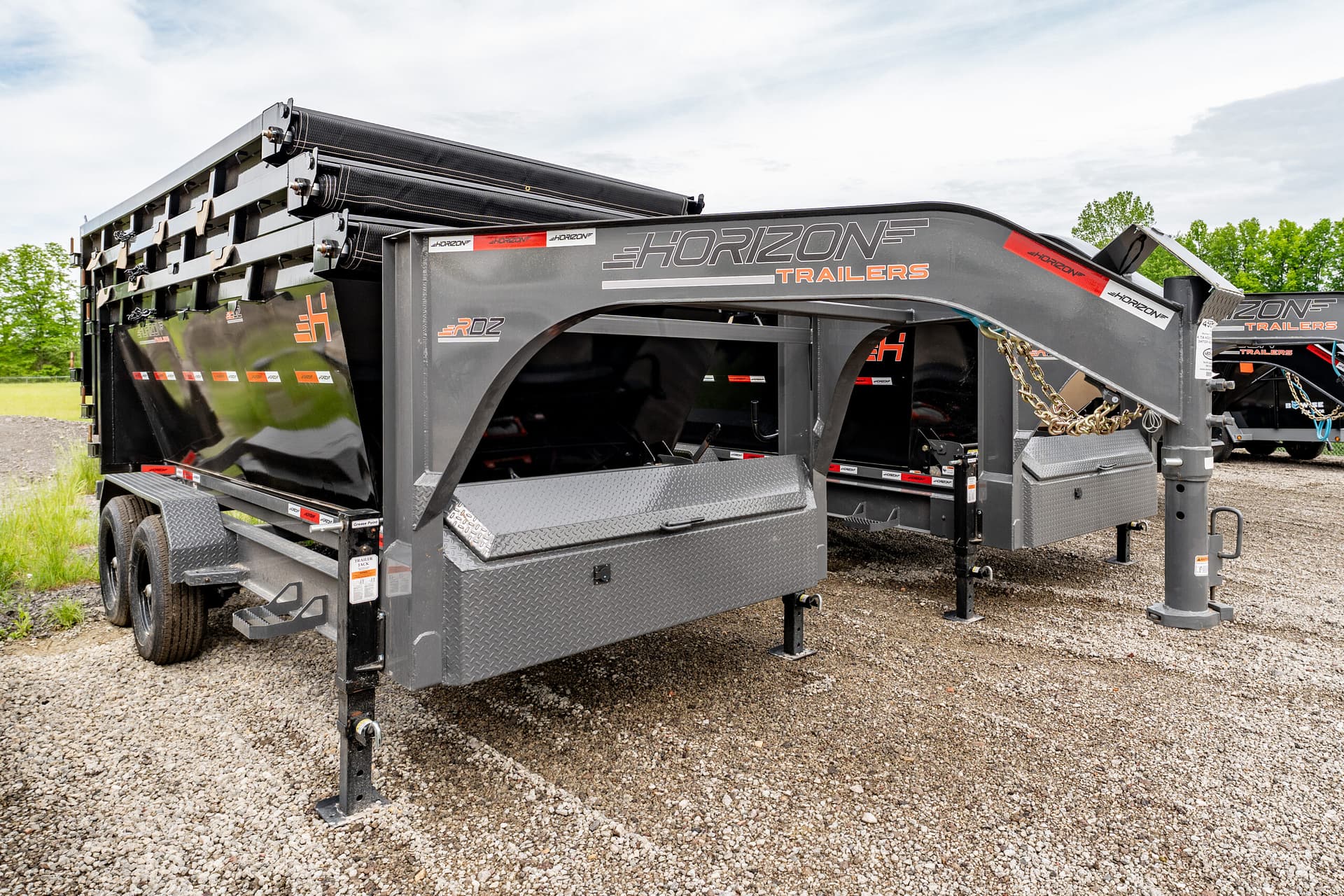 Horizon Trailers gooseneck trailer with folded loading ramps, parked on a gravel lot under a blue sky and green trees in the background.
