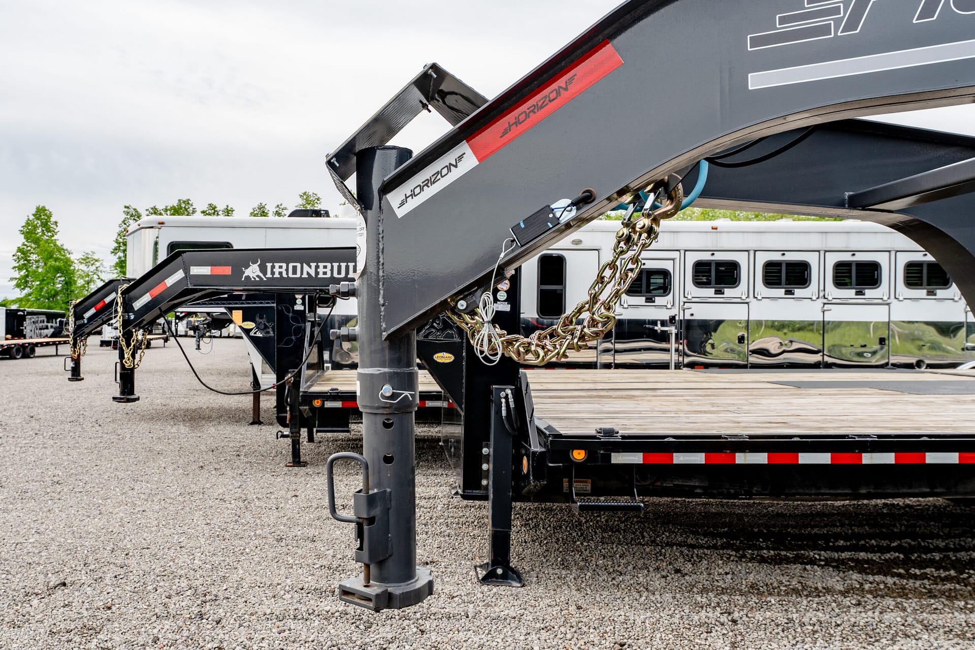 Close-up of a gooseneck trailer hitch and heavy safety chains in a gravel parking yard with trailers in the background
