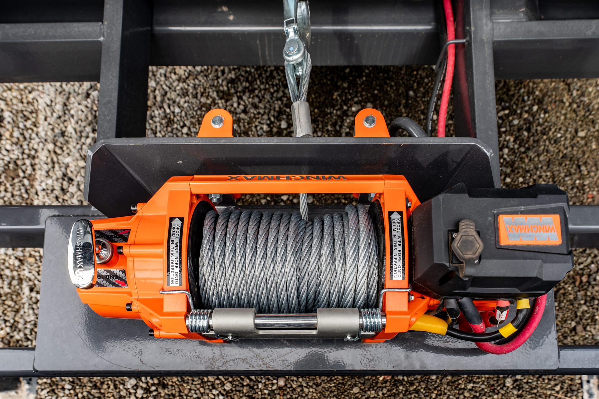 Close-up of an orange electric winch with steel cable mounted on a trailer, showing the hook and wiring on a gravel surface.