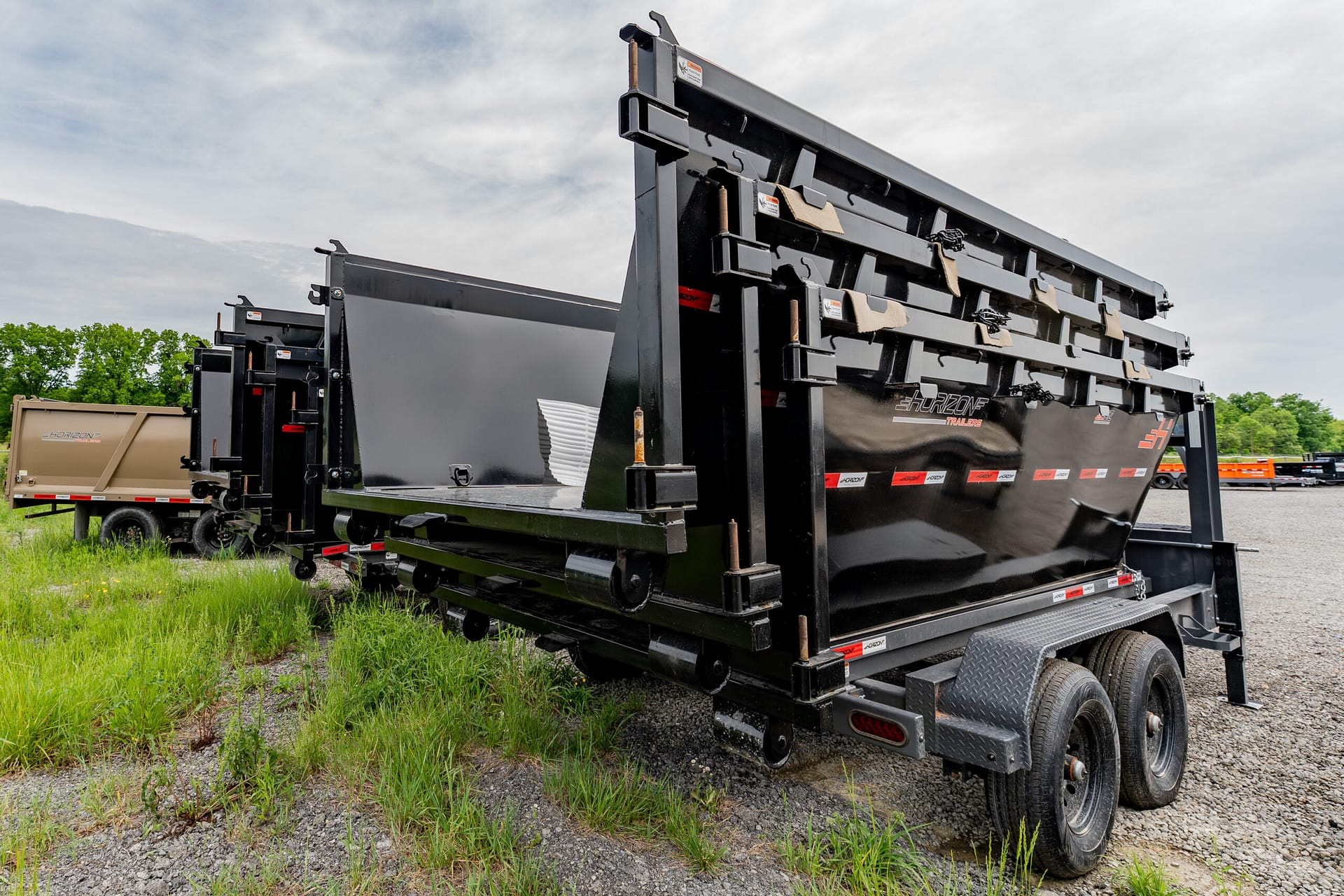 Large black dump trailer on a dual-axle truck trailer, parked on a gravel lot with a beige trailer and green trees in the background under a cloudy sky.