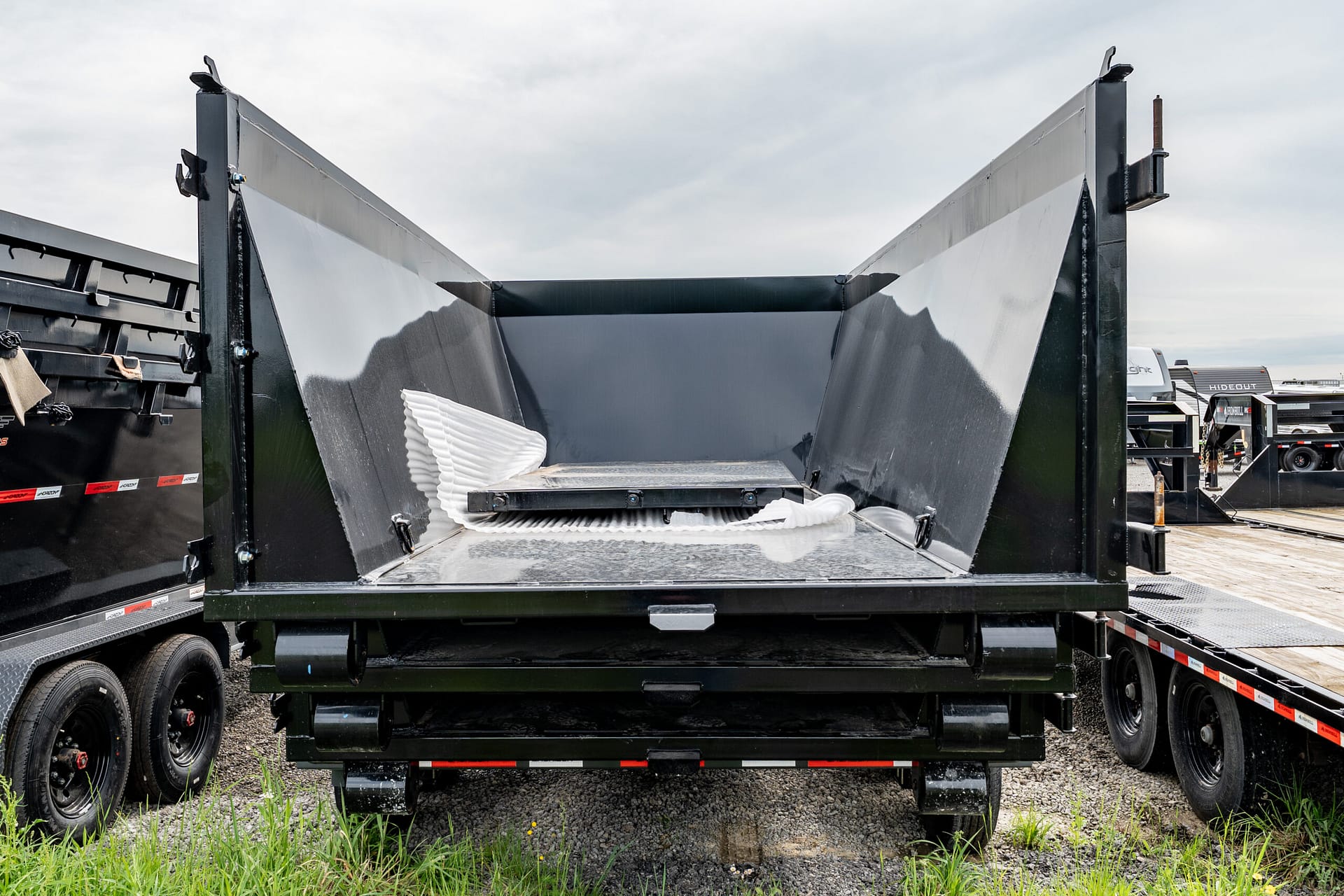Rear of a black dump trailer with its bed raised, showing the metal sides and a white corrugated hose on the bed; other trailers in a truck-yard in the background.